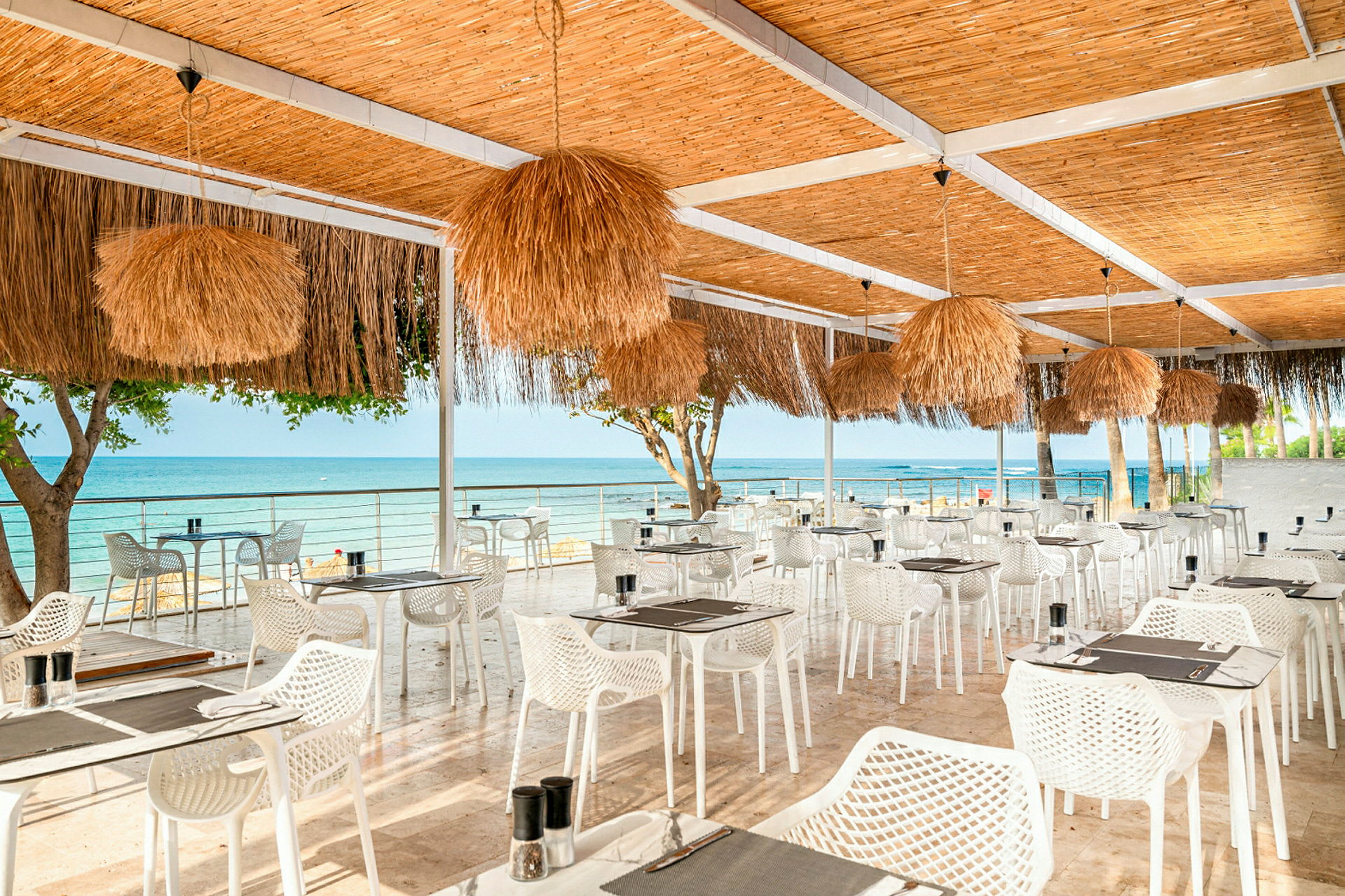 En restaurant med utendørs servering og utsikt over havet, møblert med hvite stoler og bord samt et tak dekorert med strålamper. (AI-generert tekst)