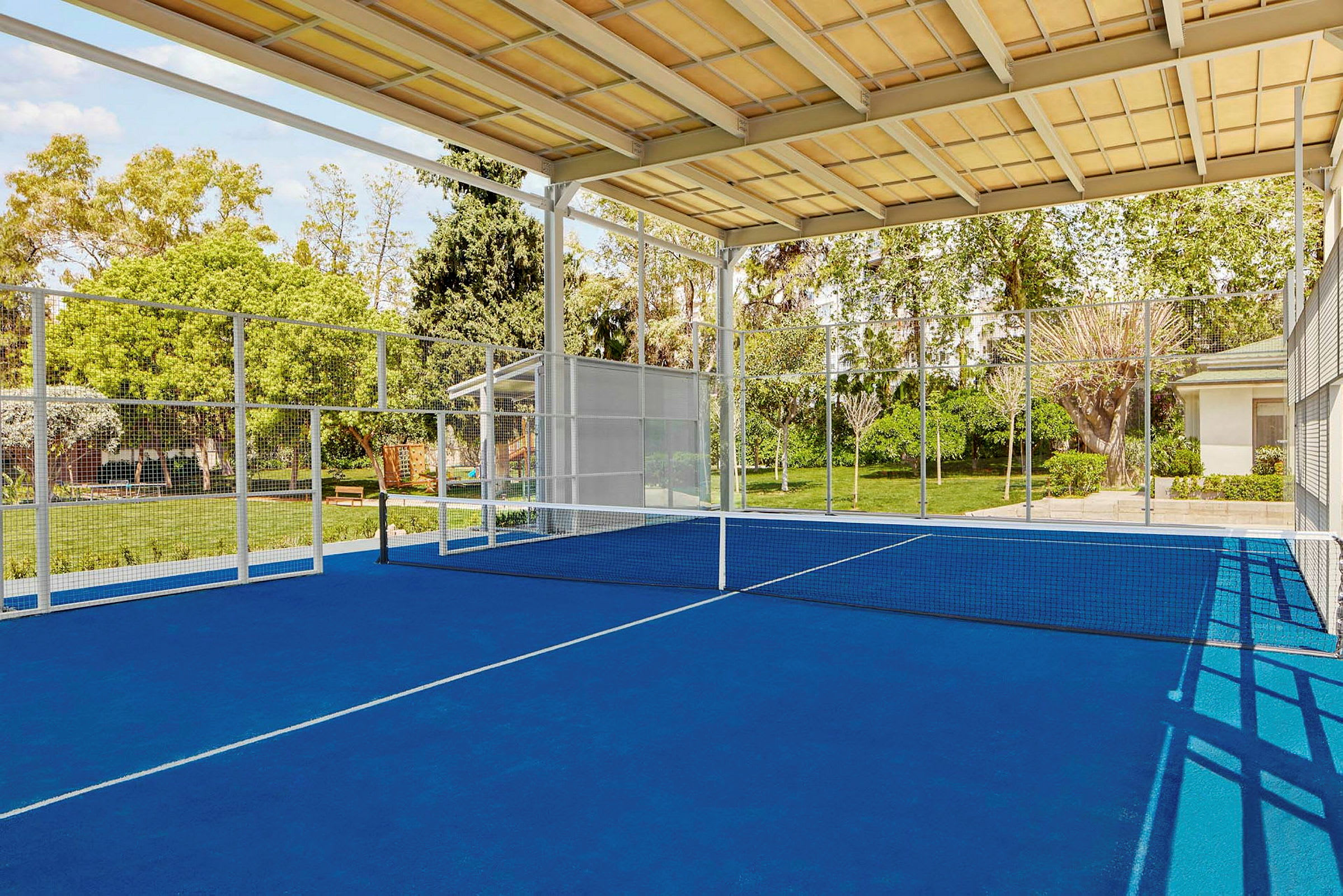 En padelbane med blå bakke, nett og omkringliggende grøntområder. (AI-generert tekst)
