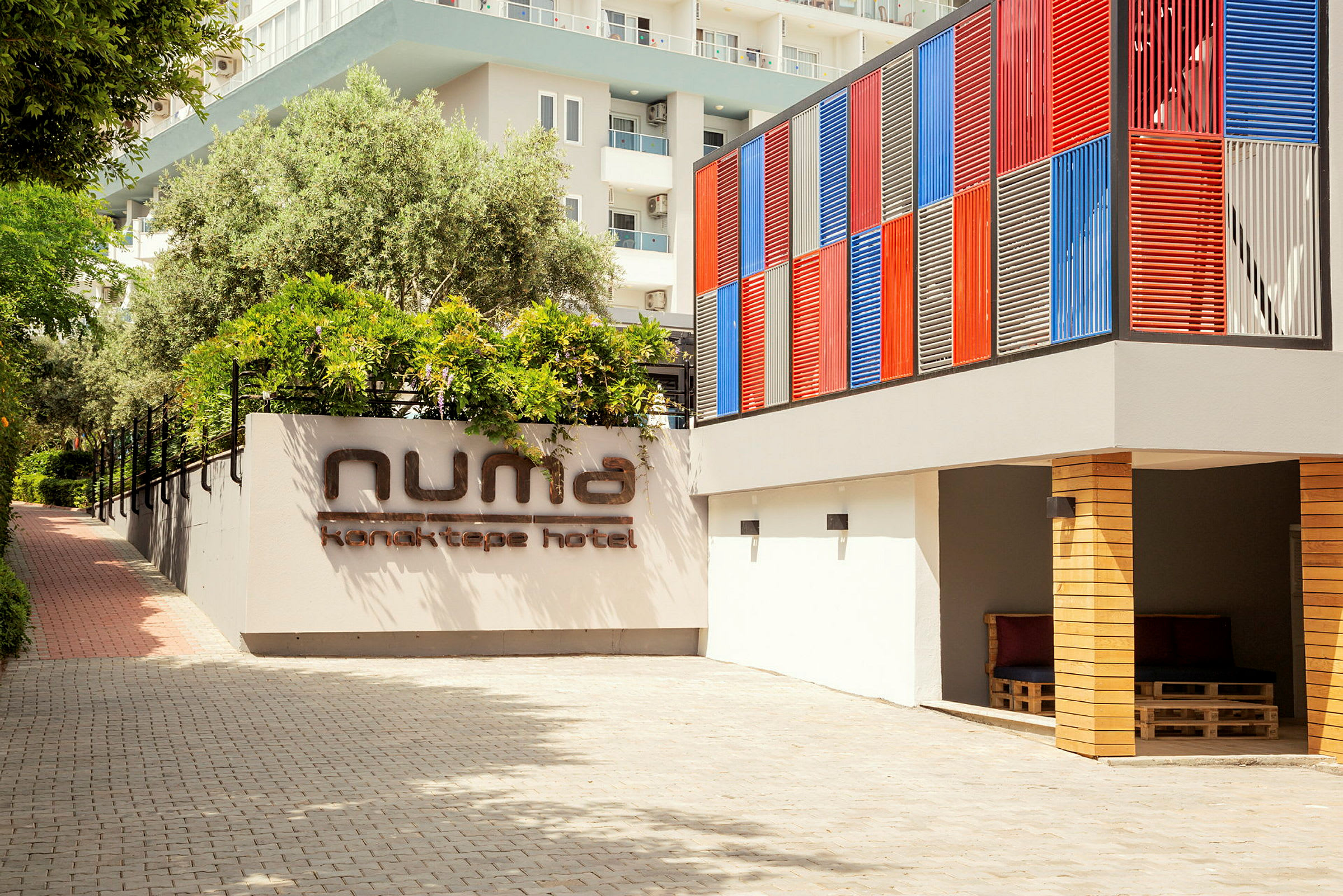 Hotellfasaden med et skilt som sier 'Numa Konaktepe Hotel', omgitt av planter og en fargerik vegg. (AI-generert tekst)