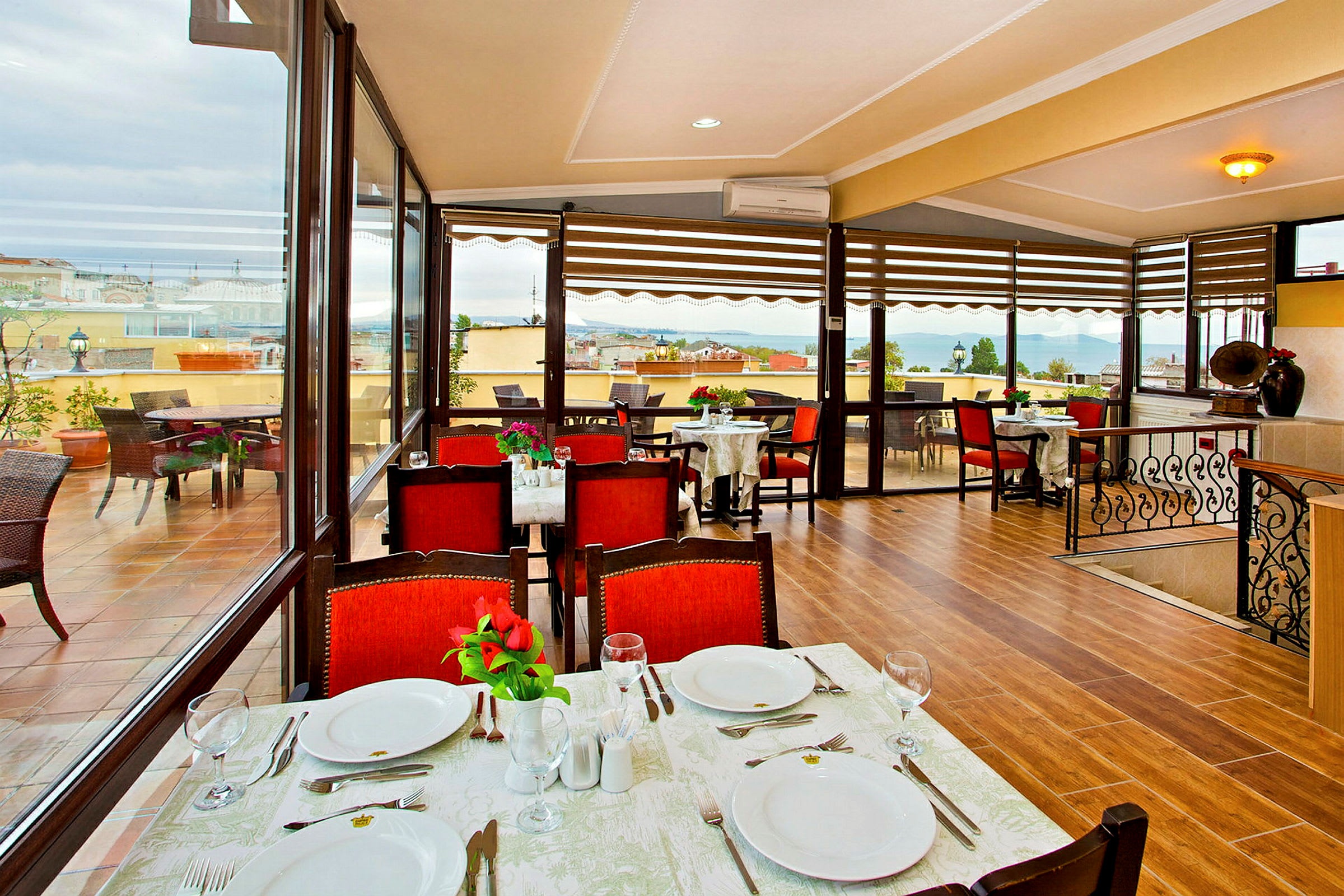 En restaurant med bord dekket for måltid, røde stoler og utsikt over en terrasse samt havet. (AI-generert tekst)