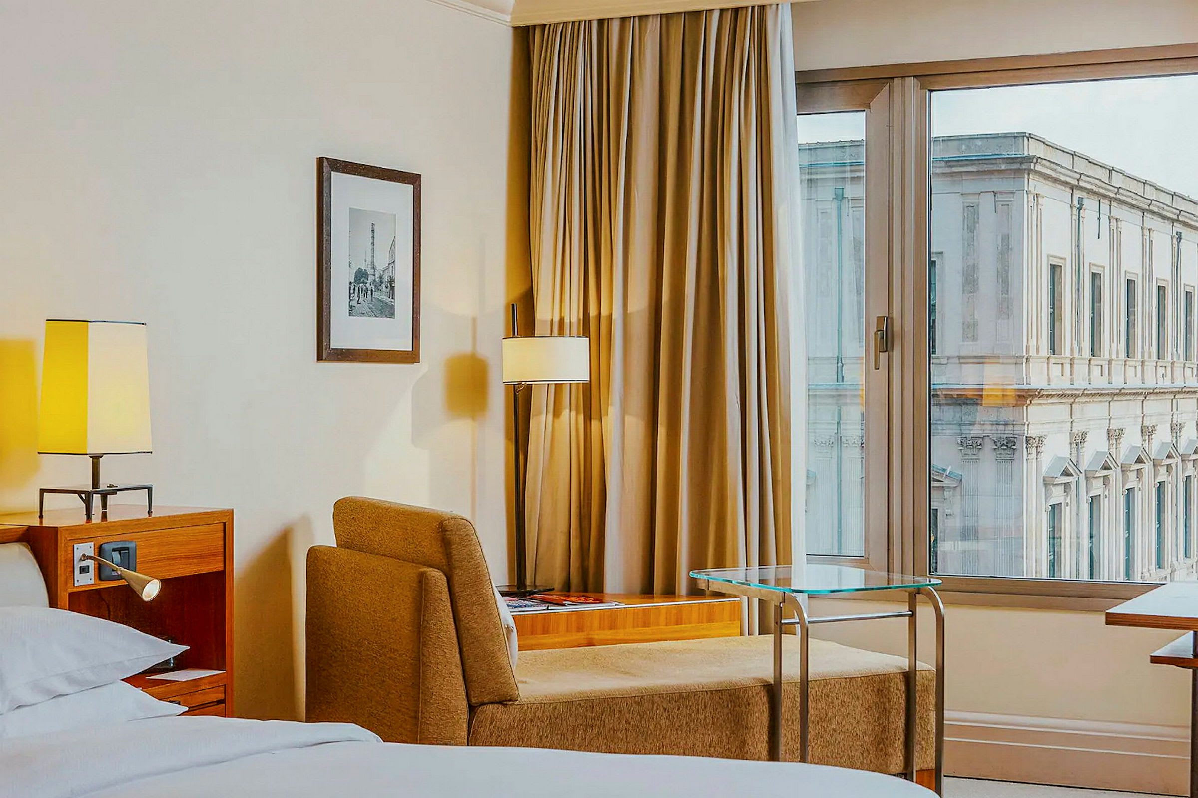 Hotellrom med seng, lenestol, bord og vindu med utsikt mot en bygning. (AI-generert tekst)