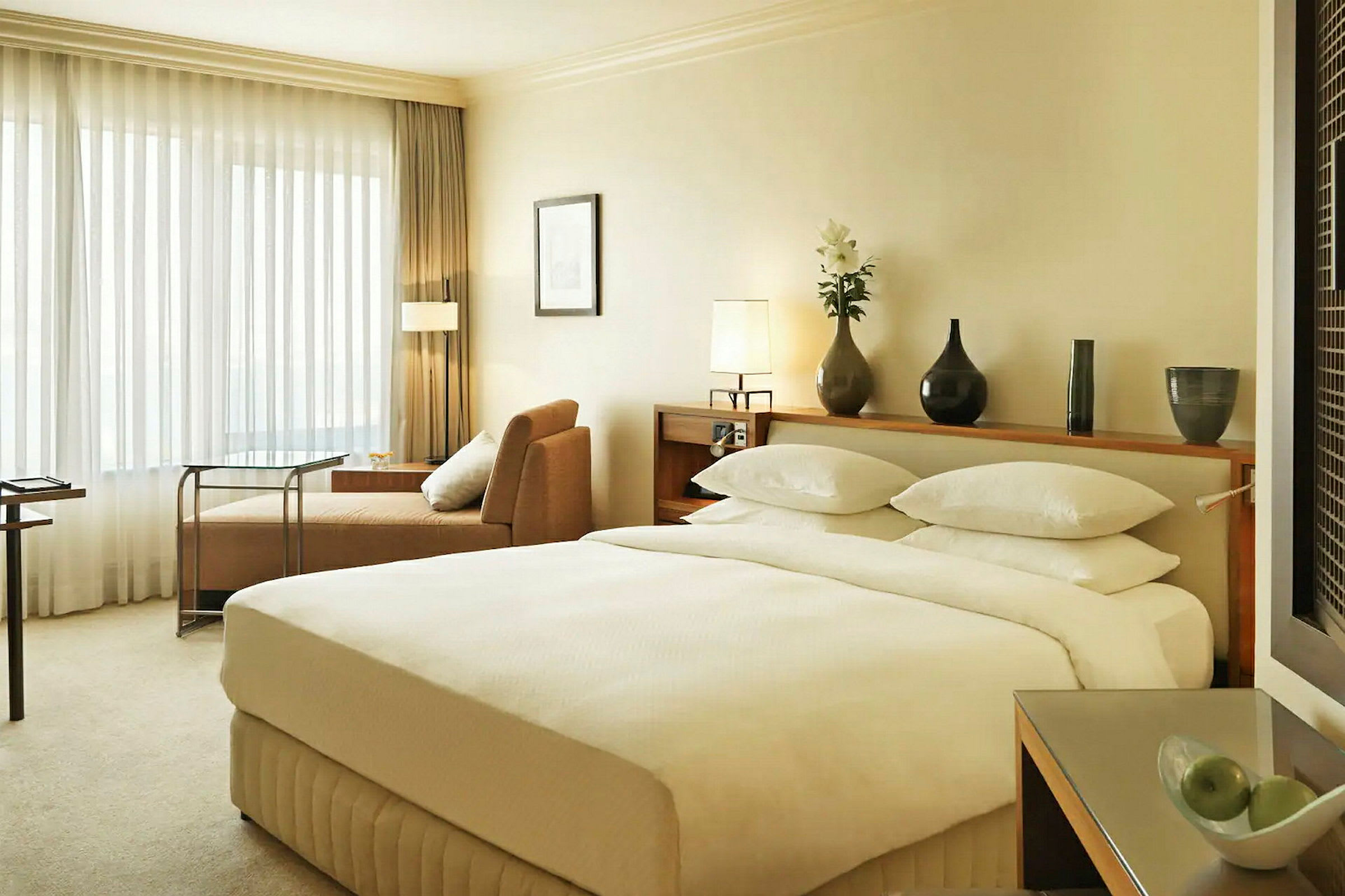 Soverom i hotellrom med en stor dobbeltseng, beige sofa og et bord nær et vindu med gjennomsiktige gardiner. (AI-generert tekst)