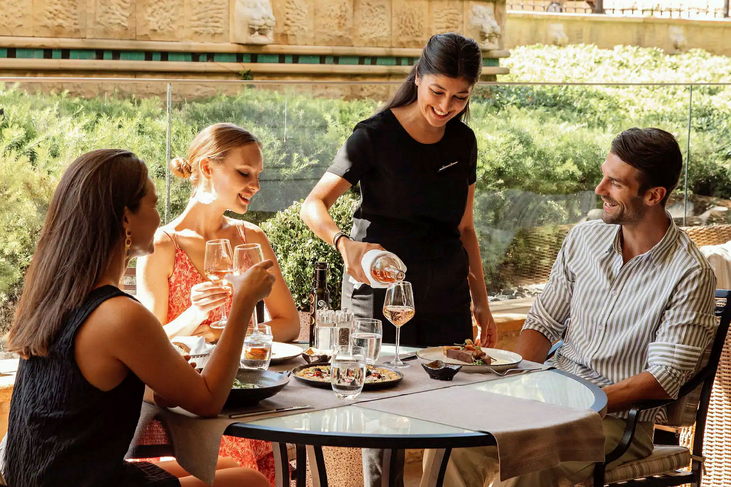En servitør serverer drikke til gjester som sitter ved et bord på en uterestaurant. (AI-generert tekst)