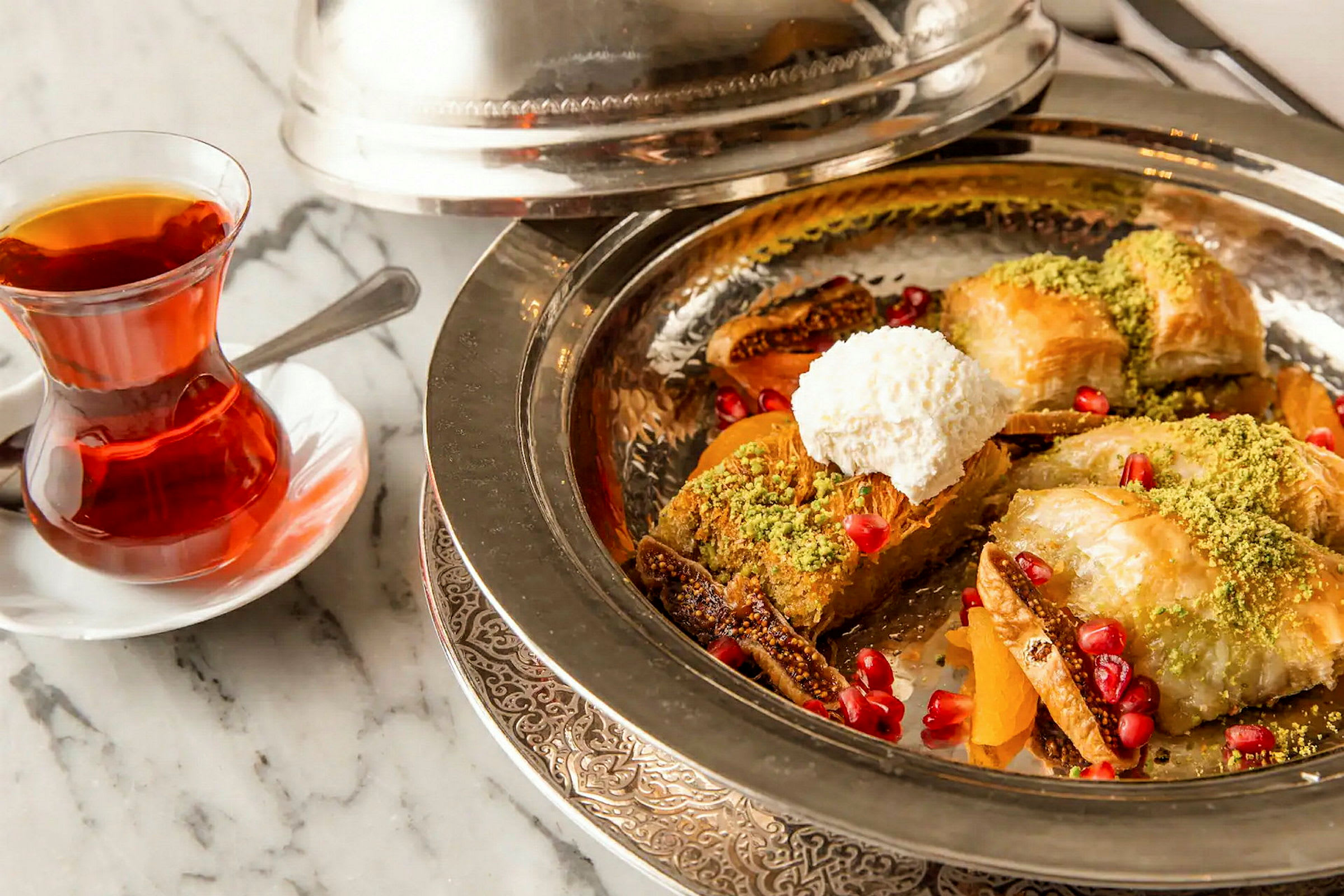 En tallerken med dessert bestående av baklava, pistasjnøtter, granateplefrø og pisket krem, ved siden av et glass te. (AI-generert tekst)