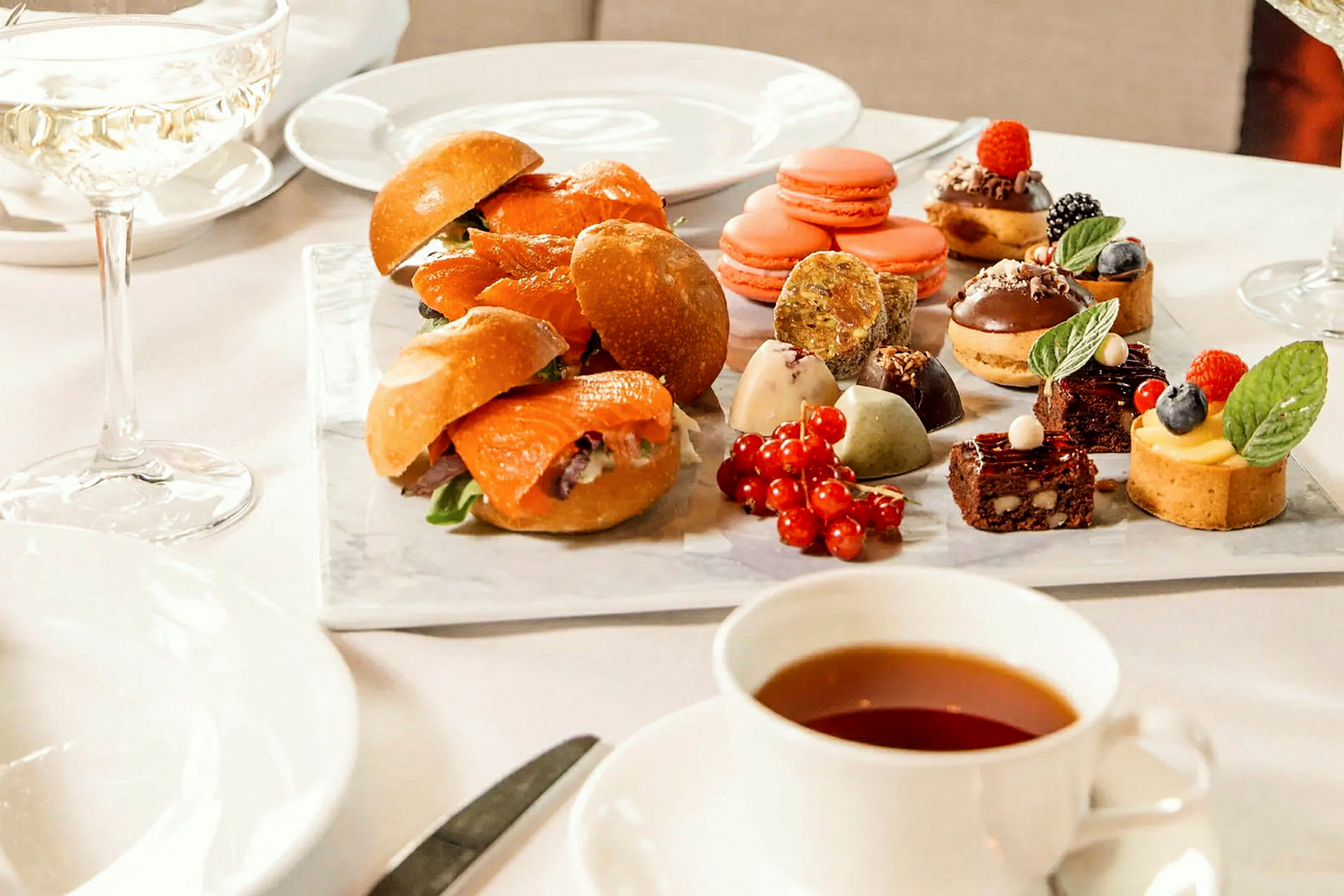 Et bord med diverse småretter og desserter, inkludert smørbrød, makroner og små bakverk. En kopp te og et glass vin er også satt frem. (AI-generert tekst)