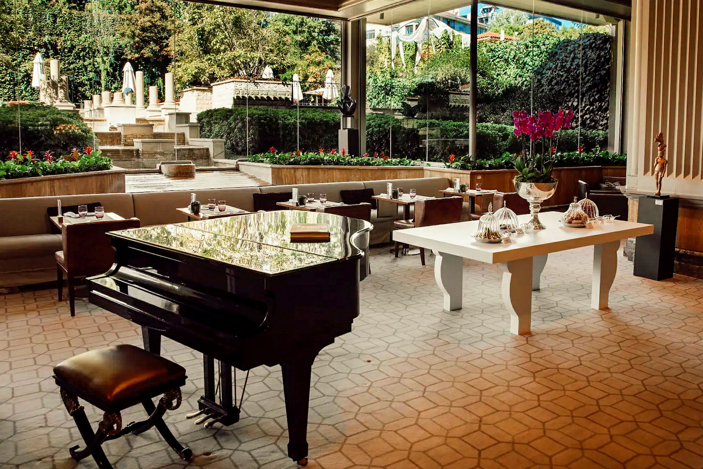 En hotellrestaurant med et stort svart piano i forgrunnen. I bakgrunnen er det sofaer, bord og utsikt over en hage med vegetasjon og trapper. (AI-generert tekst)