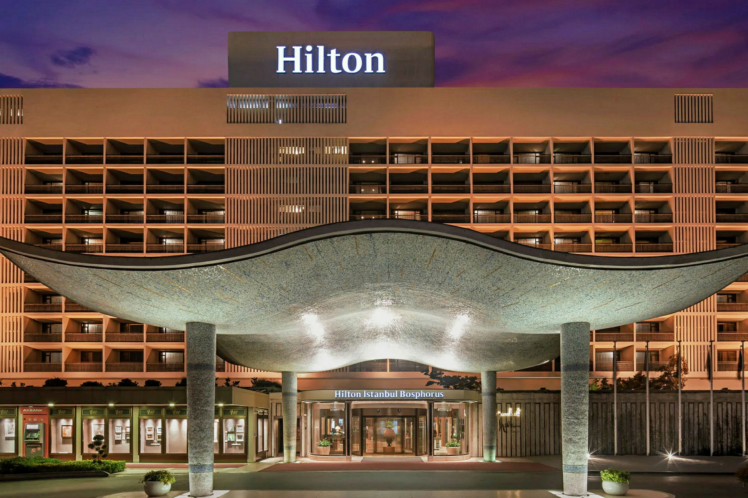Hotellfasaden med inngang under et buet tak, lyktestolper og en opplyst skilt med teksten 'Hilton'. (AI-generert tekst)