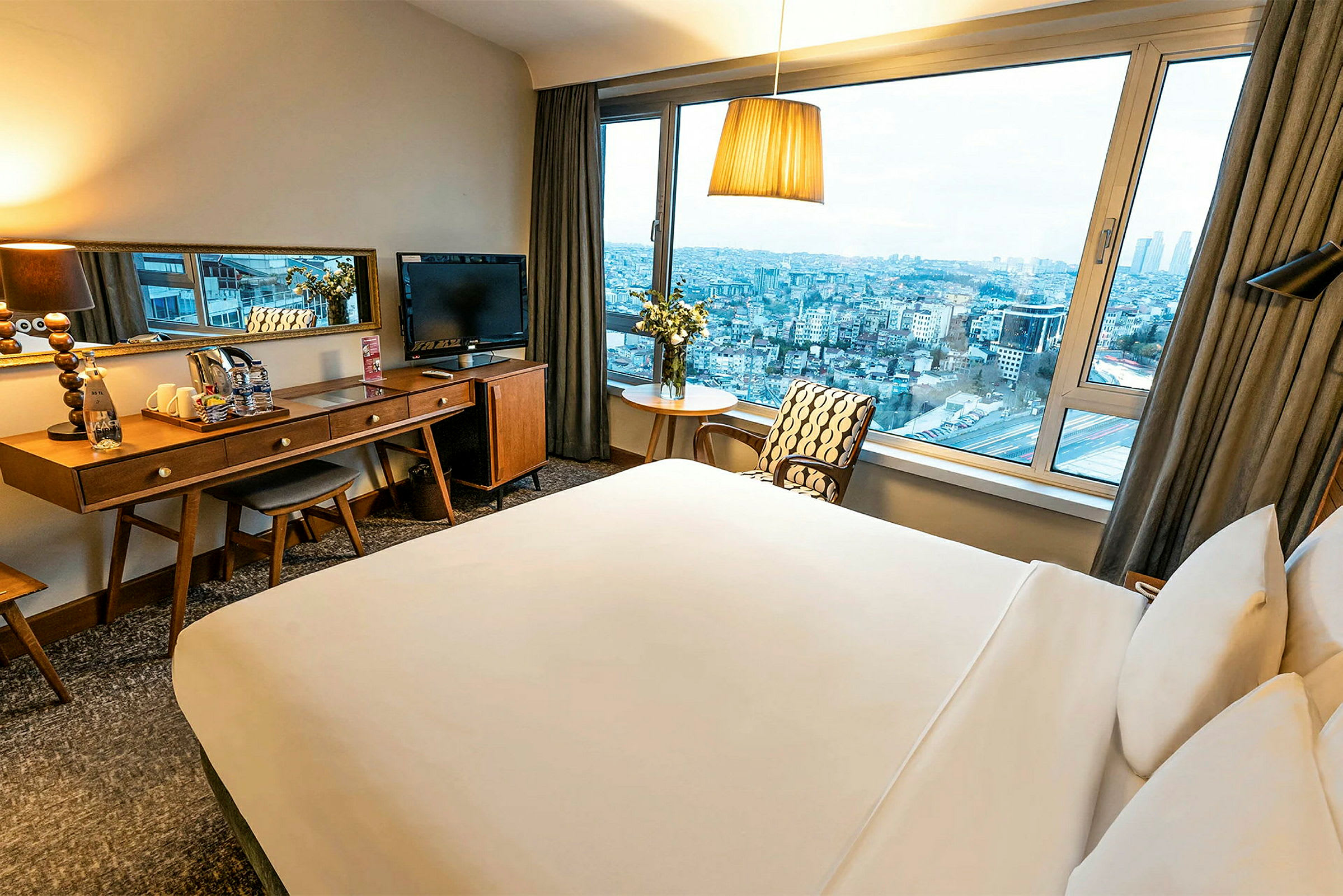 Et hotellrom med en stor seng, skrivebord og speil samt utsikt over en by gjennom et stort vindu. (AI-generert tekst)