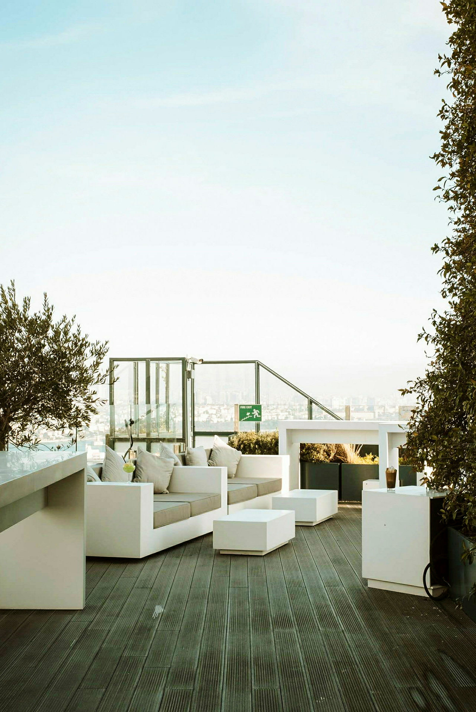 En takterrasse med moderne hvite sittemøbler og bord, omgitt av planter og med utsikt over et bylandskap. (AI-generert tekst)