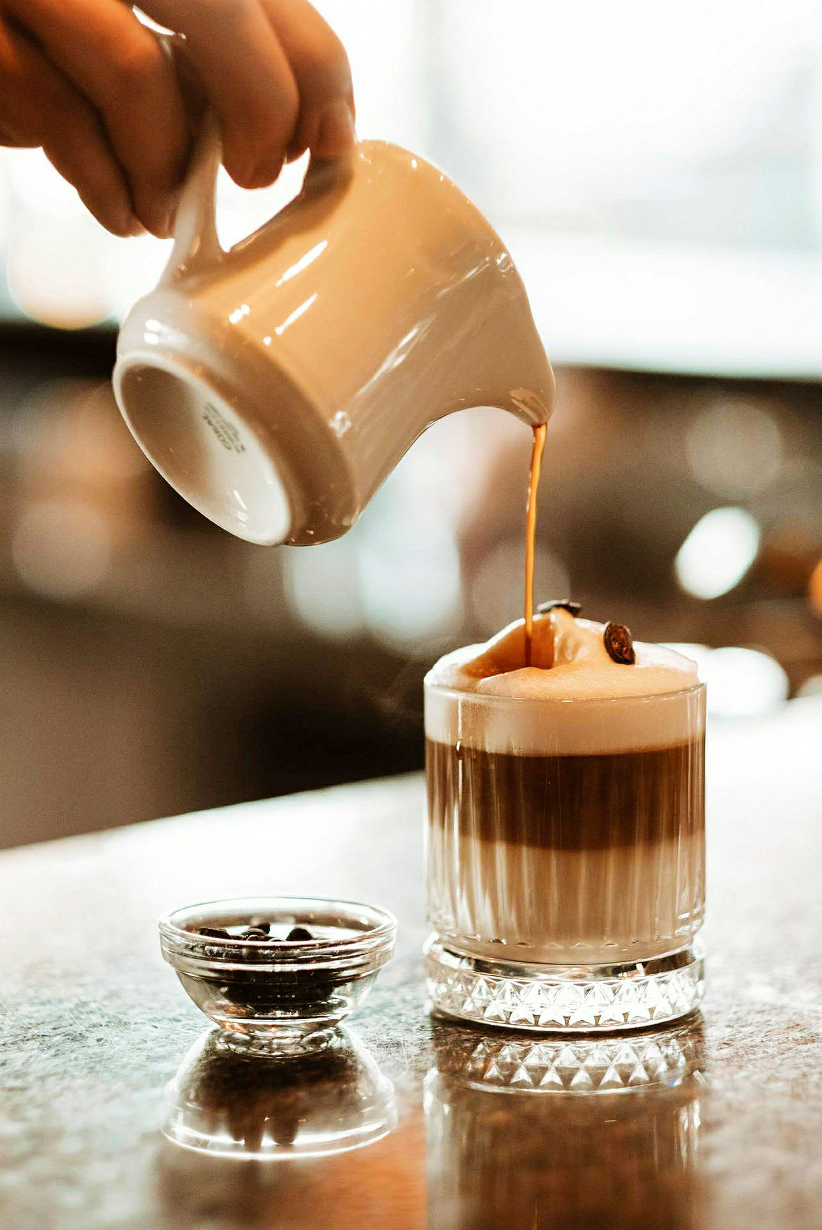 En hånd heller en varm drikk, muligens kaffe, fra en kanne ned i et glass med skum på toppen. Ved siden av glasset står en liten skål med kaffebønner. (AI-generert tekst)