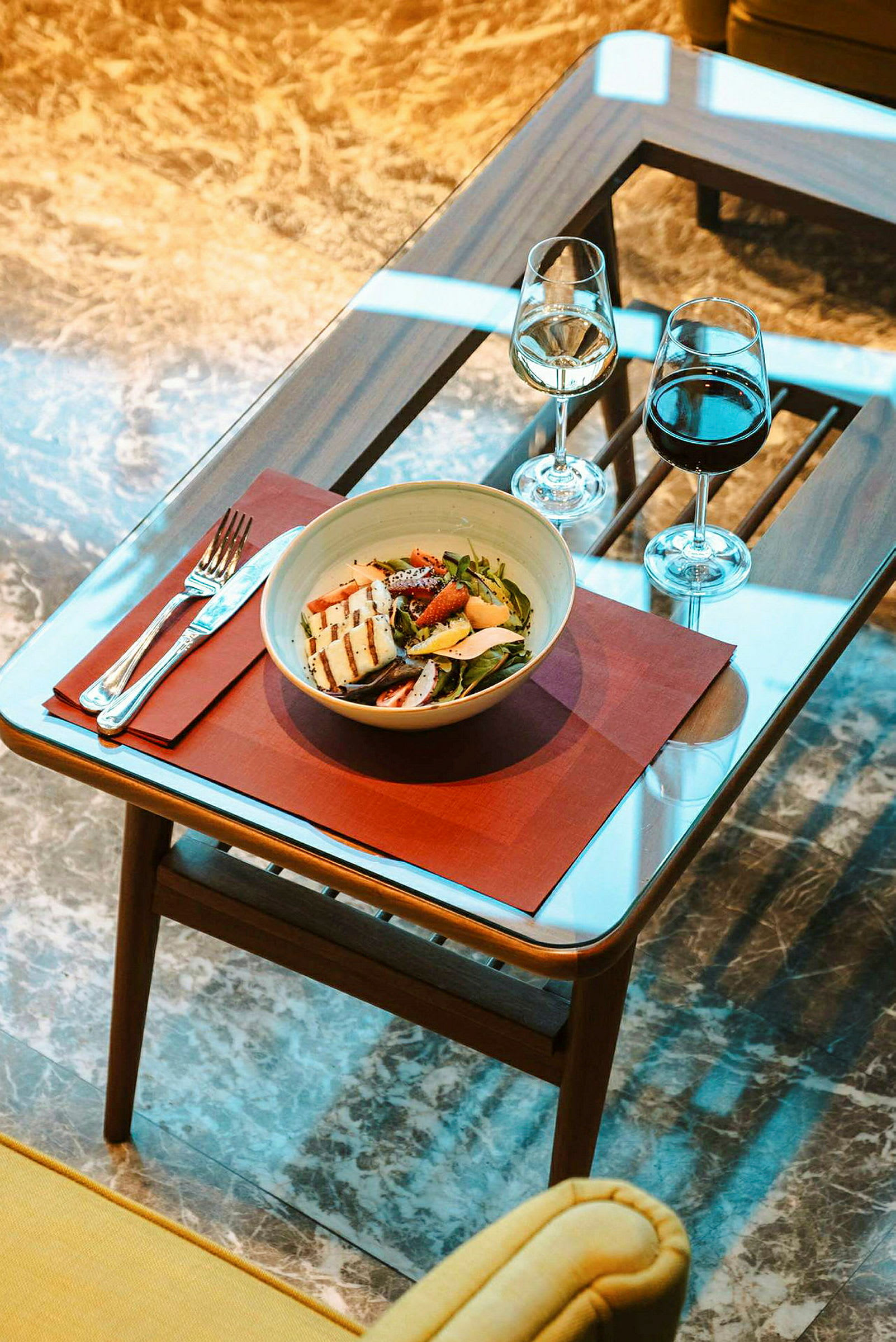Bildet viser et glassbord med en salatskål, bestikk og to vinglass plassert på en rød bordbrikke. (AI-generert tekst)