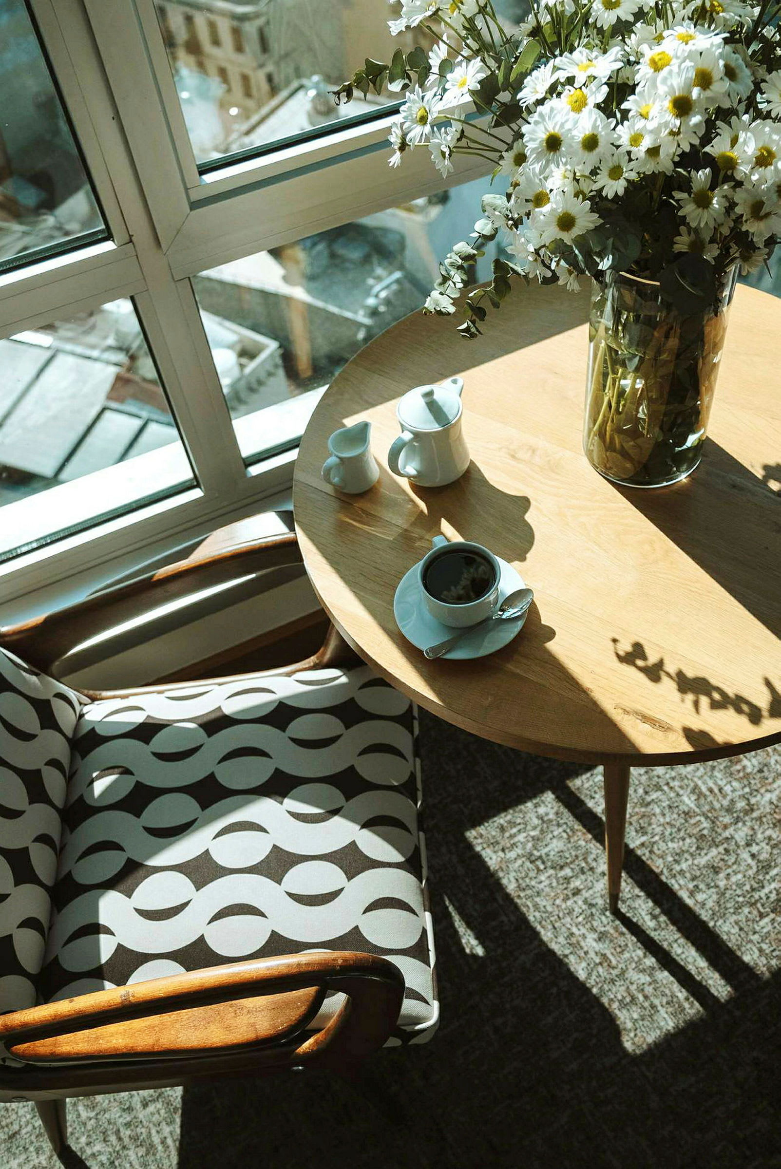 Et bord med en kaffekopp, sukkerbit og en melkekanne ved siden av en stol med mønstret stoff, sett foran et stort vindu med utsikt. (AI-generert tekst)