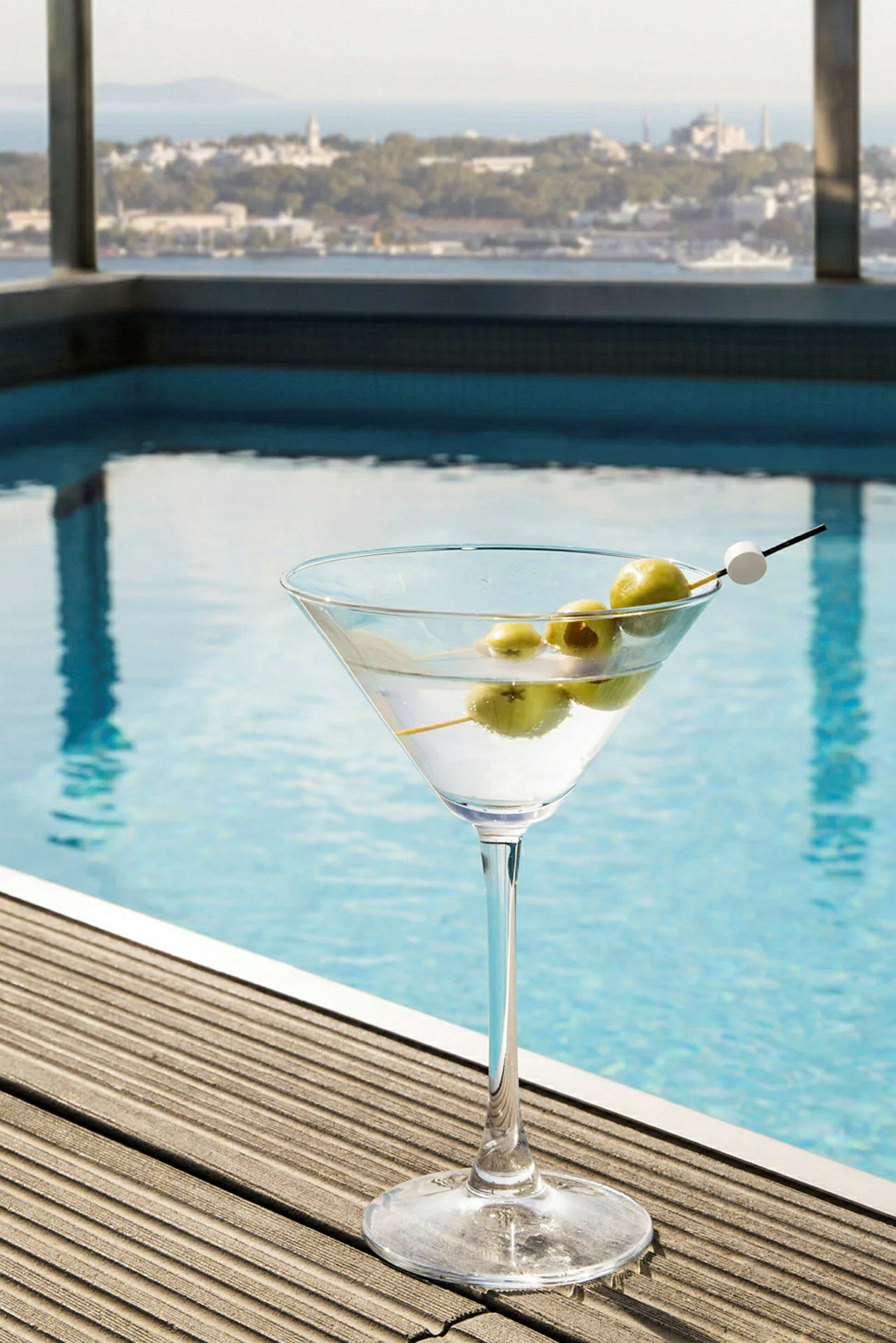 Et glass med en cocktail og oliven på en treflate ved siden av et basseng, med utsikt over et bylandskap. (AI-generert tekst)