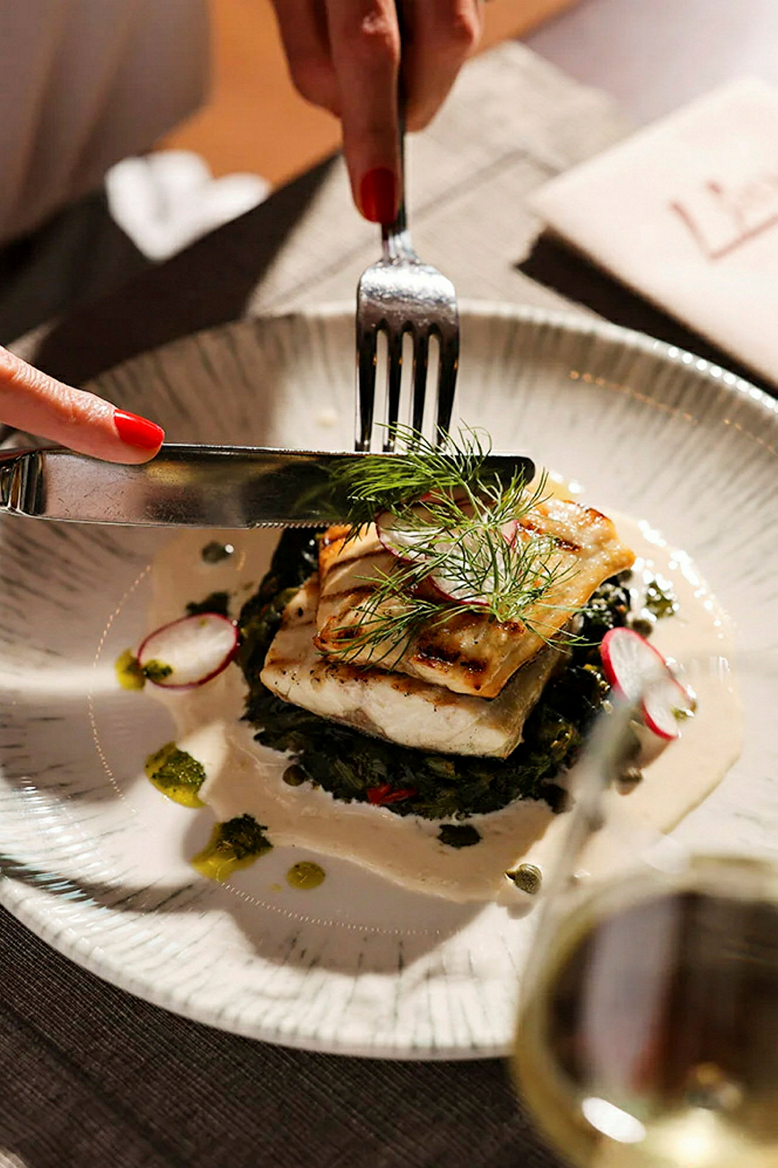 En tallerken med et måltid bestående av grillet fisk, servert med grønnsaker og en saus. Bestikk brukes på maten. (AI-generert tekst)