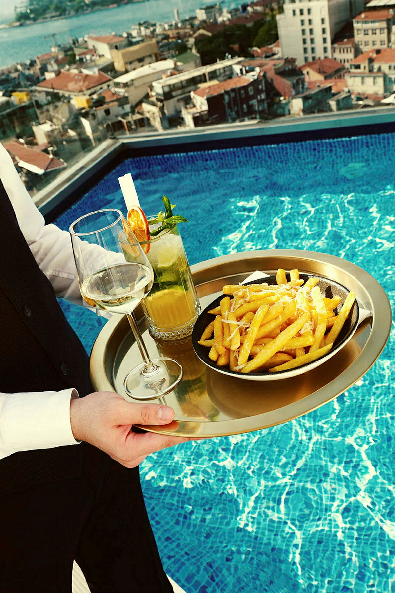 Et serveringsbrett med pommes frites på en tallerken, et glass hvitvin og en cocktail plasseres ved en basseng med utsikt over en by. (AI-generert tekst)
