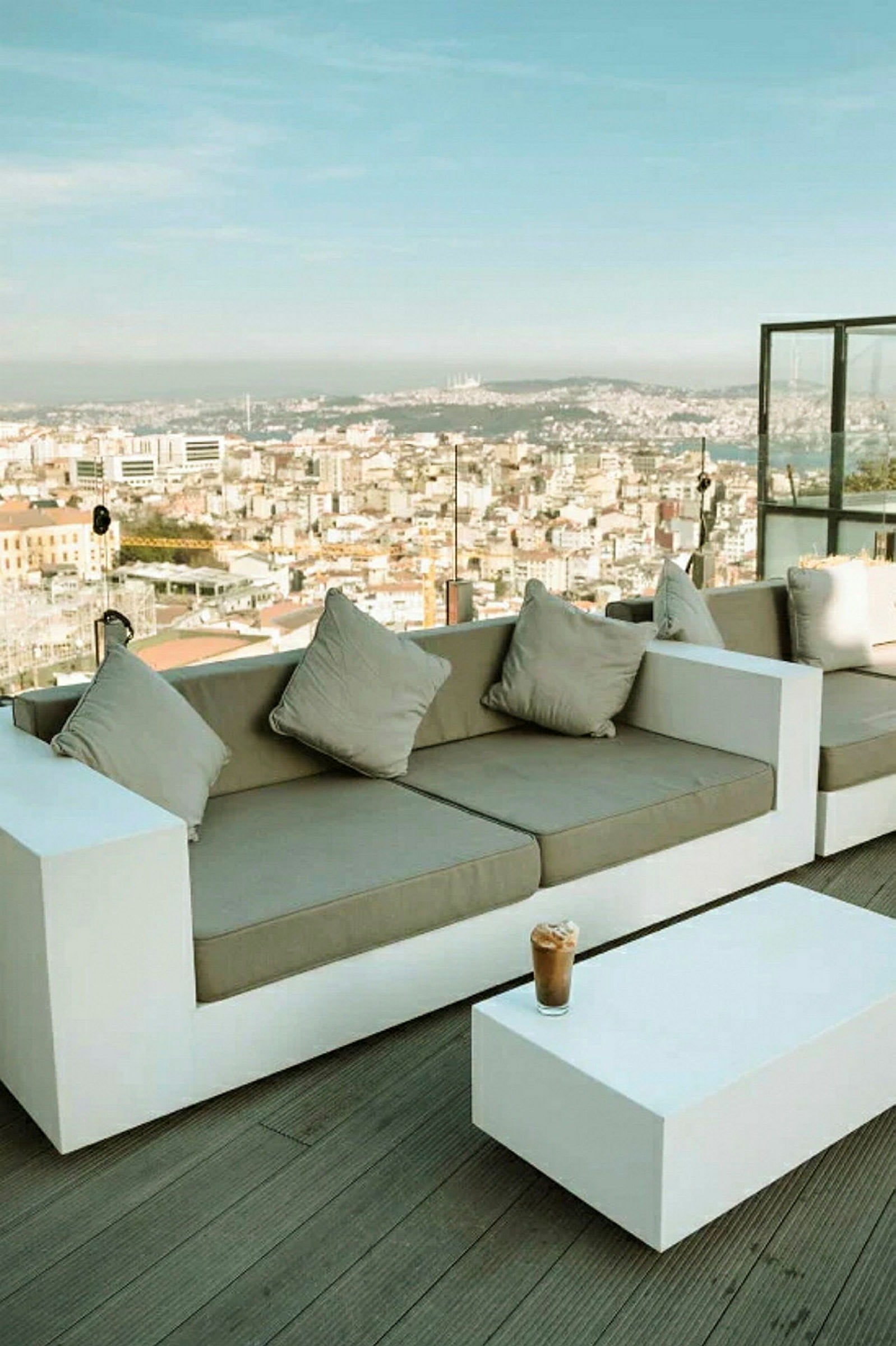 En moderne terrasse med hvite sofaer og et lavt bord, med utsikt over en by og en klar himmel. (AI-generert tekst)