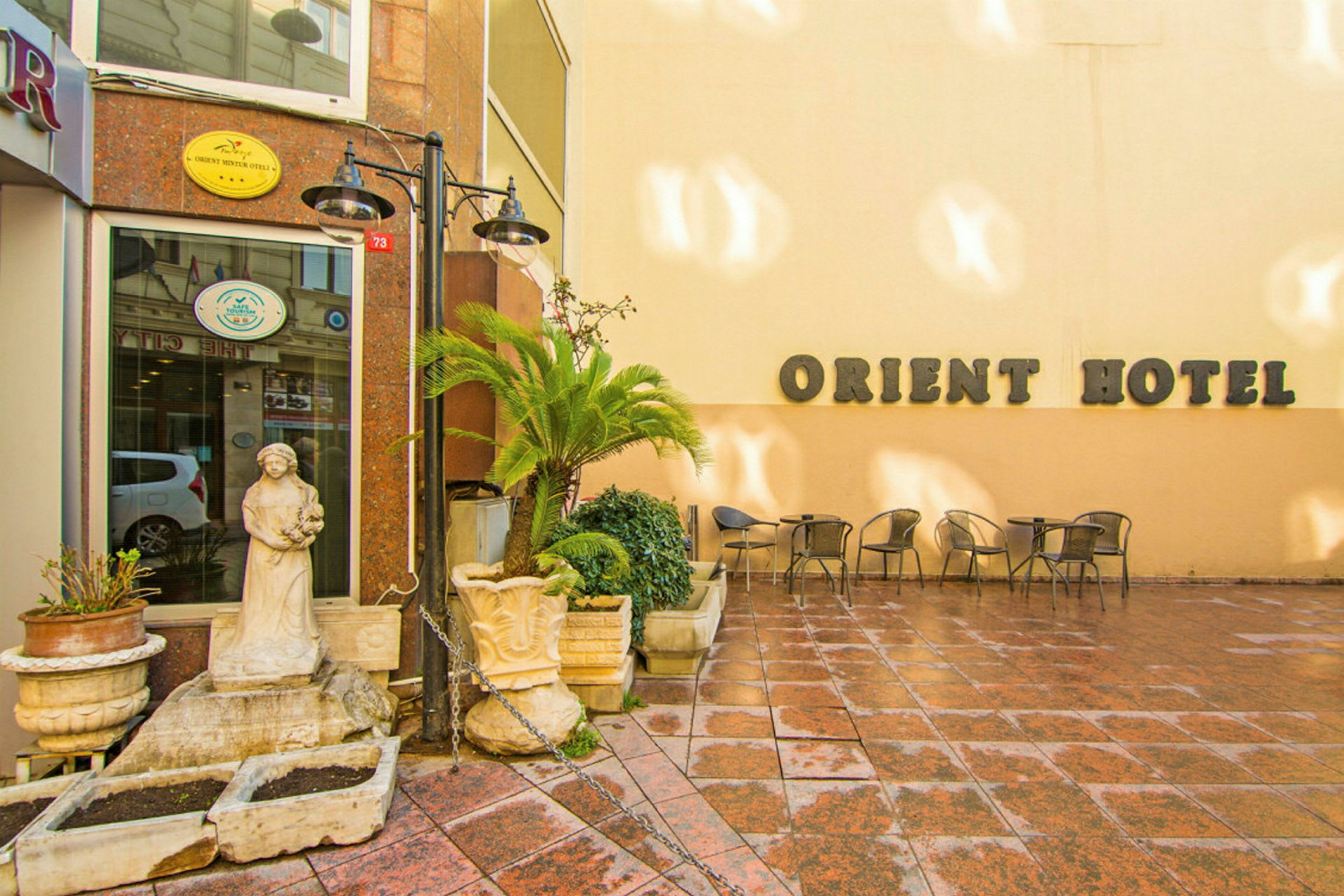 En bygning med skiltet "Orient Hotel" på en lys vegg, med en liten uteplass dekorert med stoler, bord, blomsterpotter og en statue. (AI-generert tekst)
