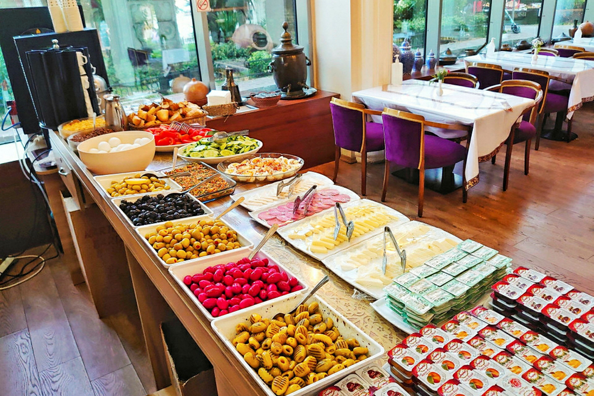 Buffet med forskjellige retter, boller og tallerkener fylt med grønnsaker, oster og pålegg. I bakgrunnen vises spiseplass med dekkede bord og stoler. (AI-generert tekst)