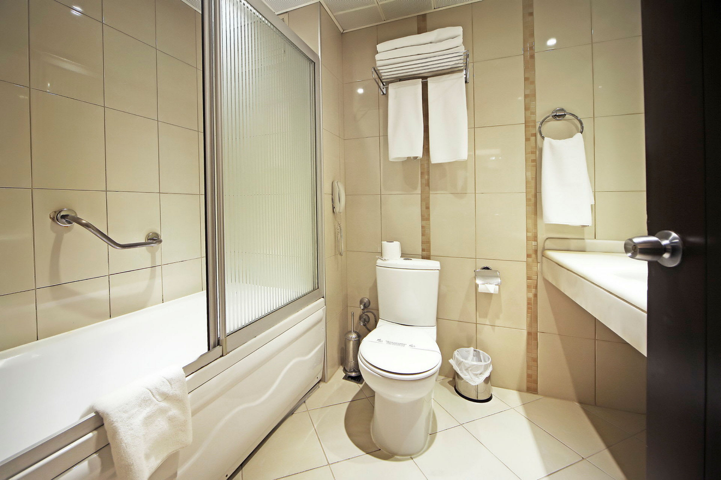 Et bad med badekar, toalett og servant. Veggene har beige fliser, og badet er utstyrt med håndklær og en papirkurv. (AI-generert tekst)