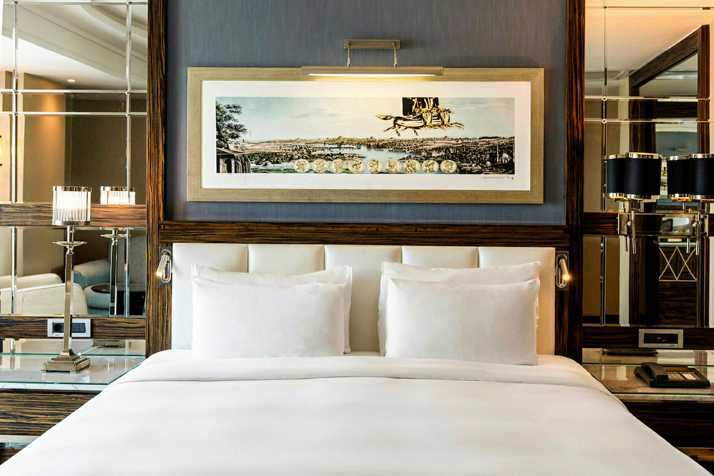 Et elegant hotellrominteriør med en bred, hvit oppredd seng foran en vegg dekorert med innrammet kunstbilde, speil og elegante lamper. (AI-generert tekst)