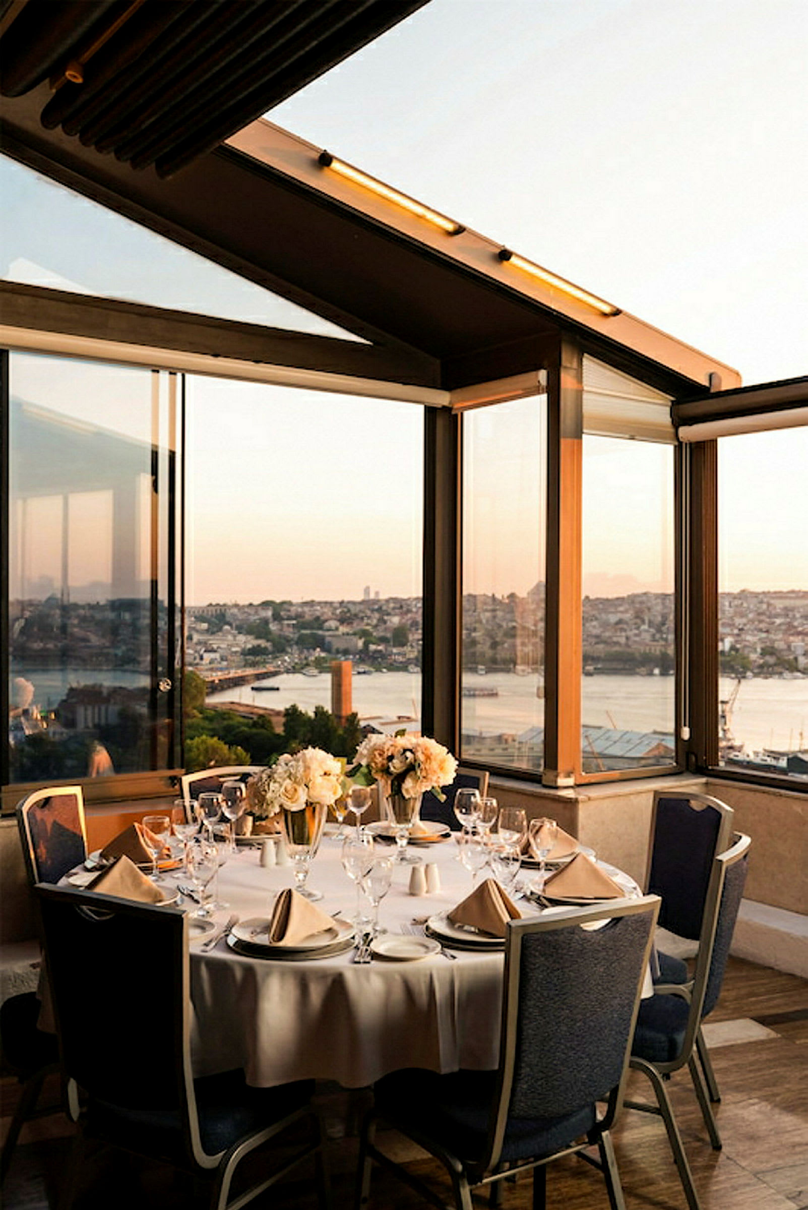En rund spiseplass med hvitt duk og elegante oppsett med servise og glass, plassert i et moderne innglasset miljø med utsikt over en by og en vannvei ved solnedgang. (AI-generert tekst)