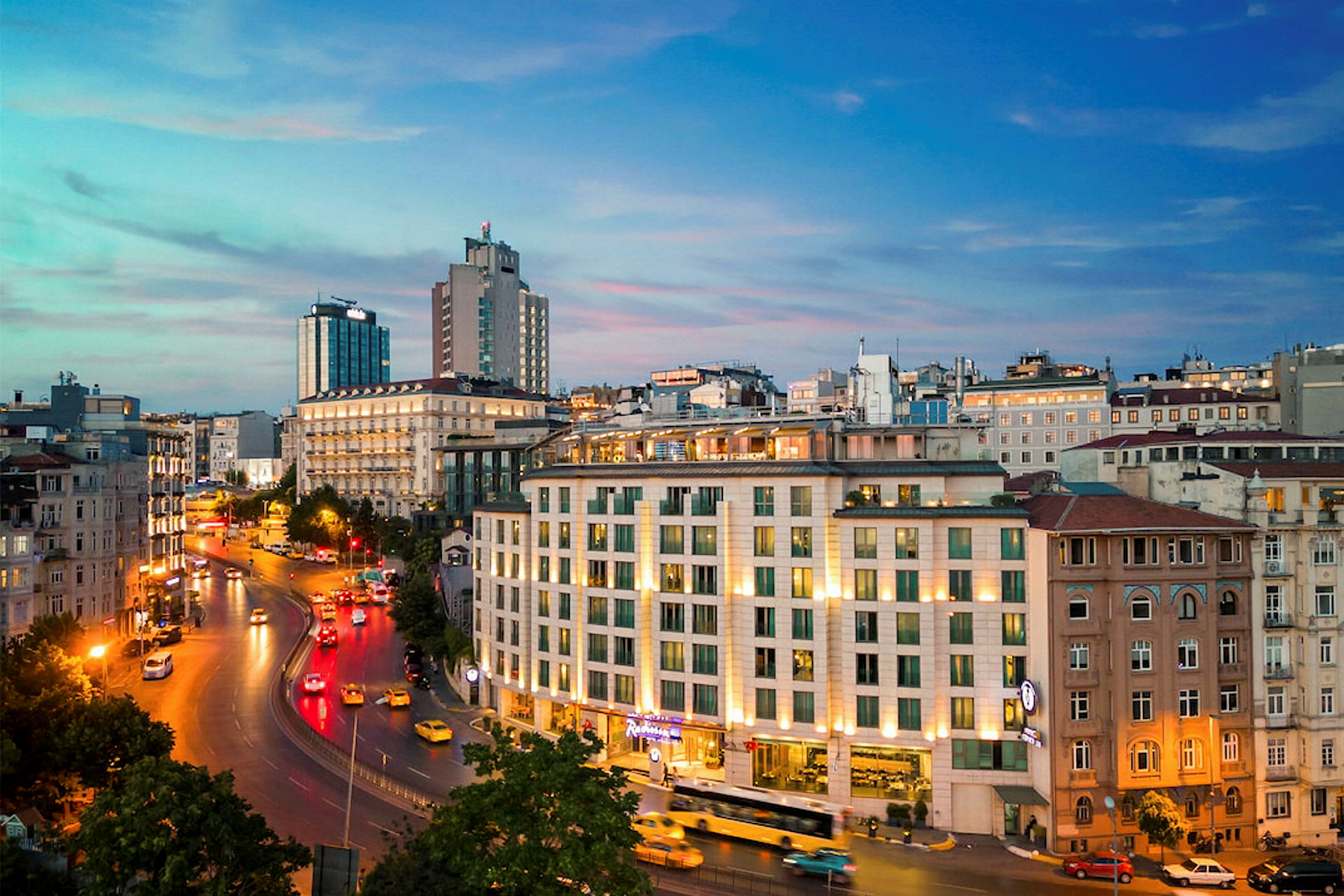 Panoramabilde av et bymiljø i skumringen med en vei, bygninger og lys fra biler. (AI-generert tekst)