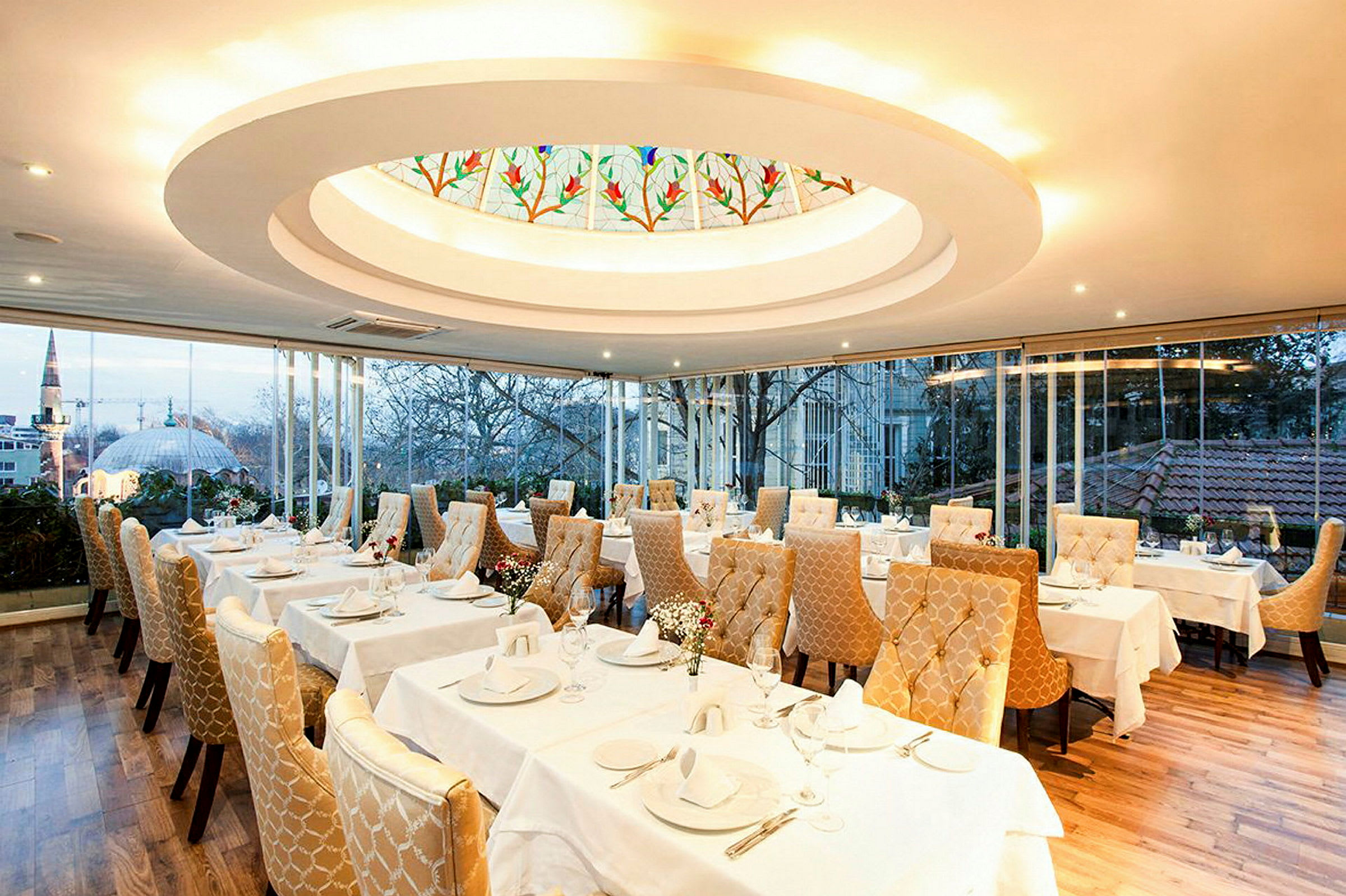 En elegant restaurant med oppdekkede bord og gyldne polstrede stoler. Et stort vindu gir utsikt over en by med trær og bygninger. (AI-generert tekst)