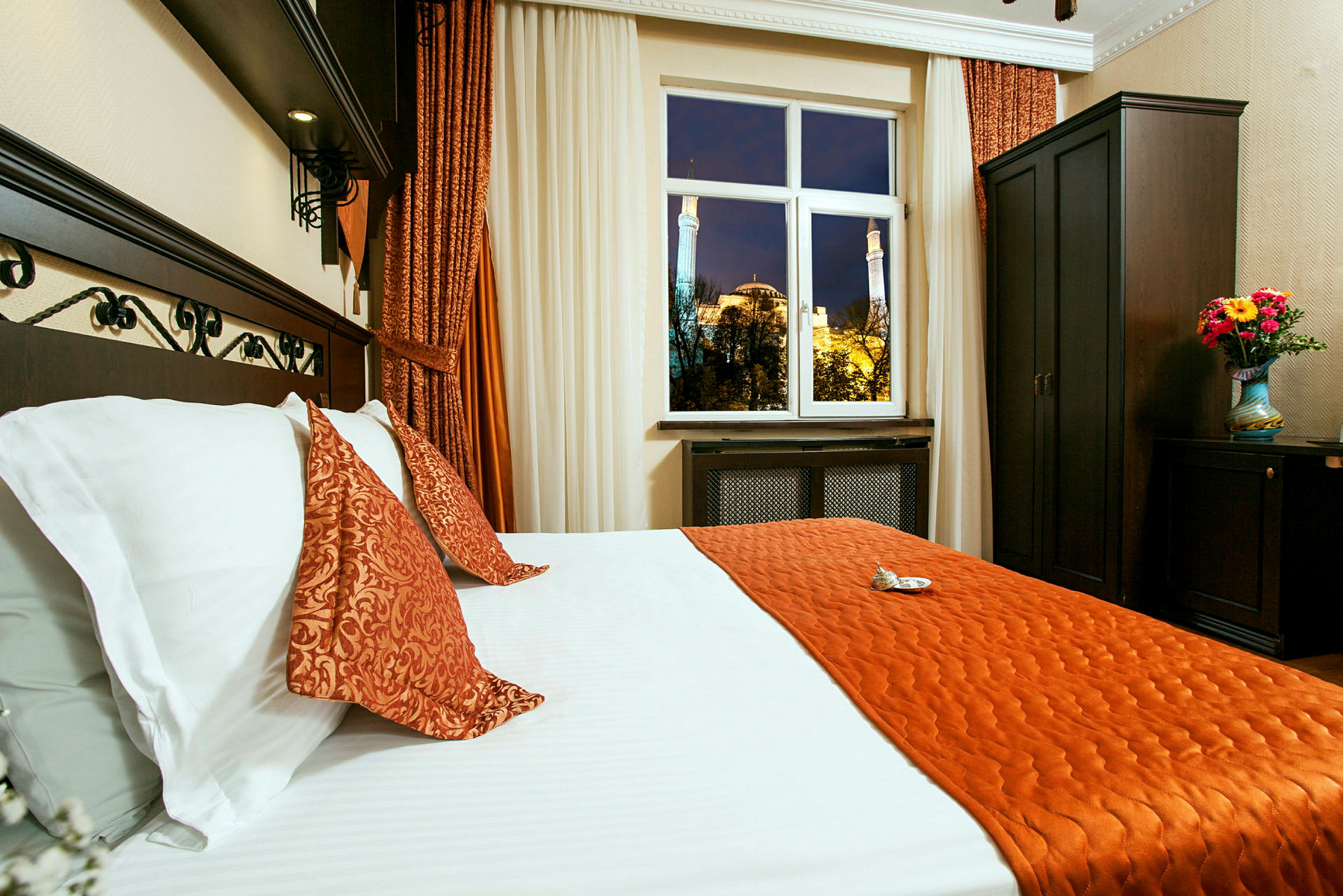 Et hotellrom med en seng kledd i hvite og oransje sengetøy. Et vindu viser en kveldsvy av Hagia Sofia utenfor. Mørke møbler og en vase med blomster sees i rommet. (AI-generert tekst)
