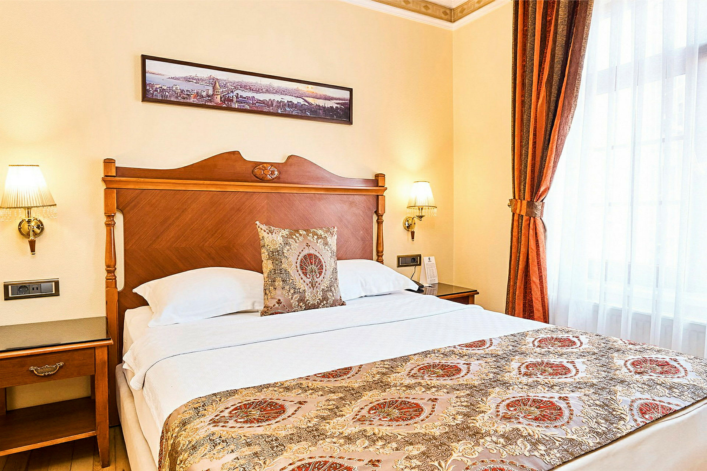 Hotellrom med en dobbelseng og elegante dekorasjoner. Sengegavlen er i tre, og sengen har et dekorert sengeteppe samt pynteputer. På veggen over henger et panoramabilde som kunst. (AI-generert tekst)
