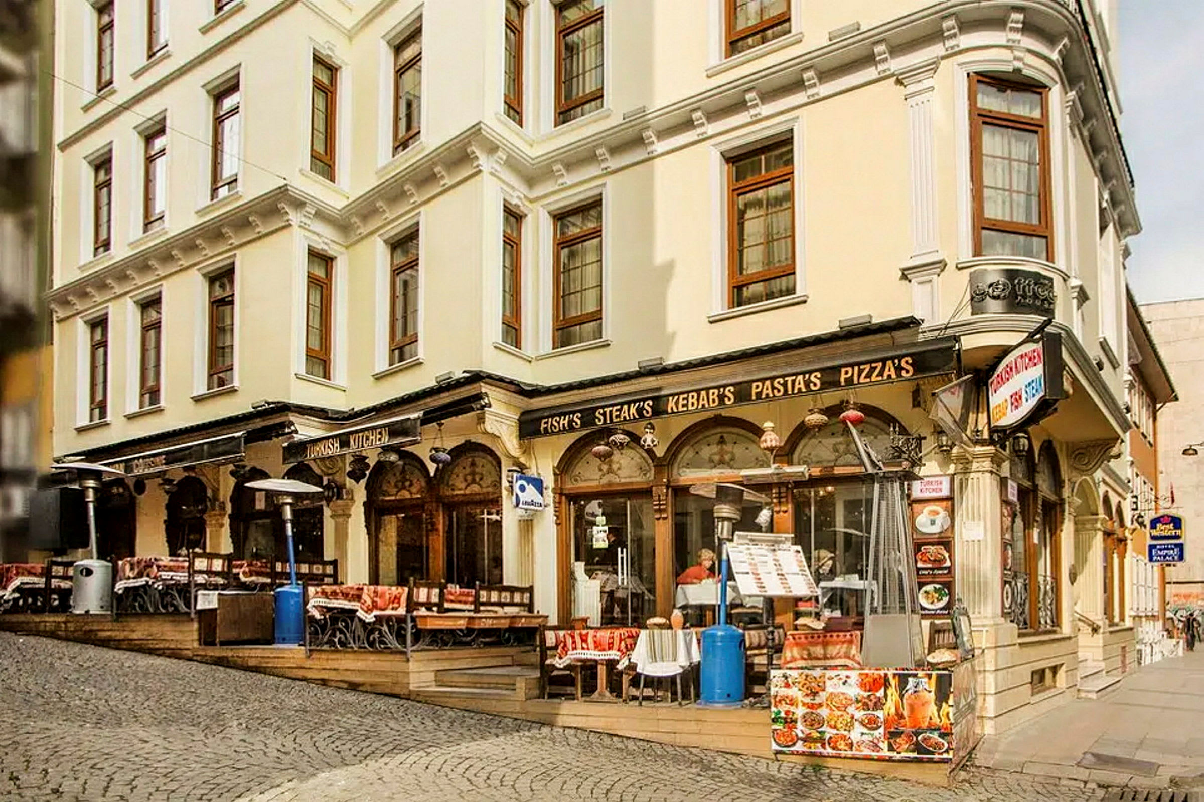 En gate med en bygning som har restaurant med uteservering, skilt med mat som kebab og pizza. (AI-generert tekst)