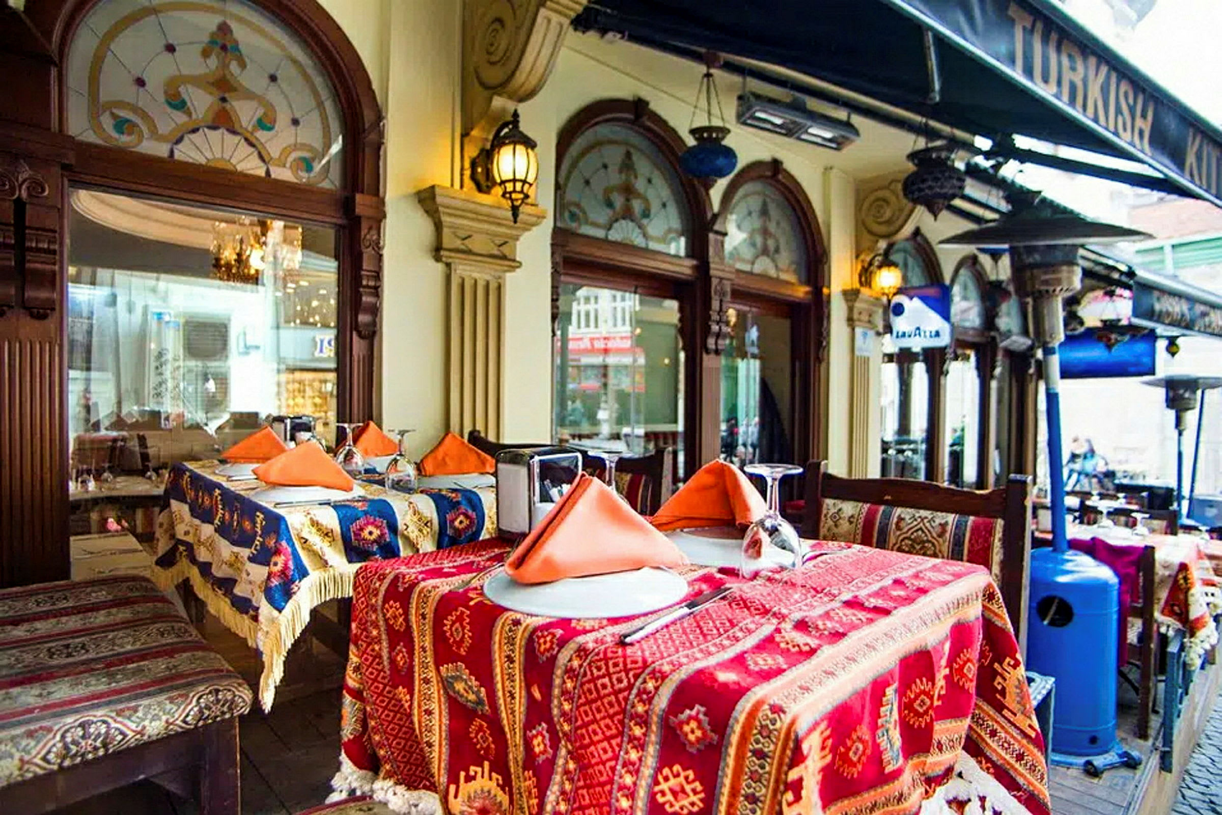 Bord dekket med fargerike duker og oransje servietter utenfor en restaurant med dekorerte vinduer og tak under en baldakin. (AI-generert tekst)