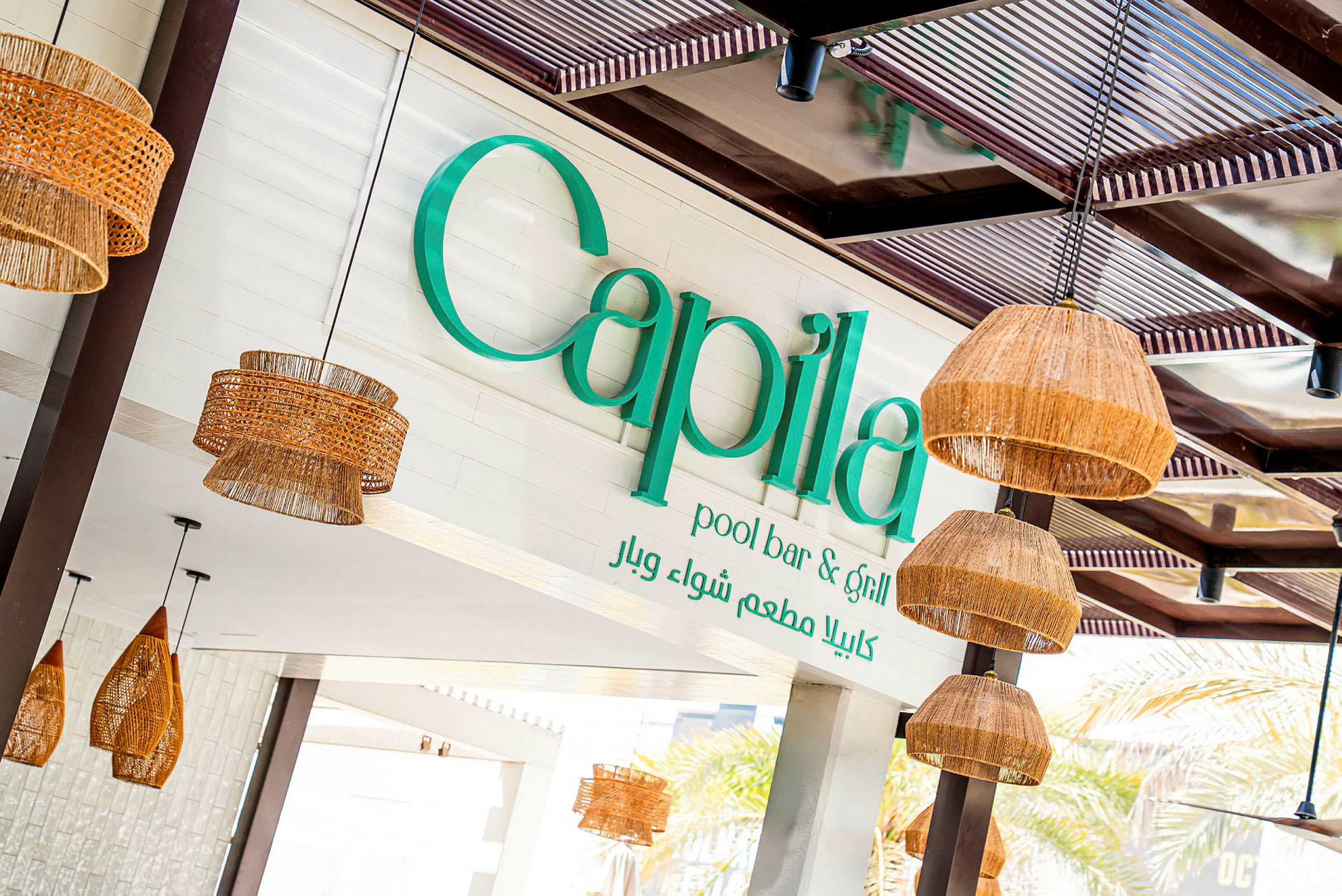 Et utendørsskilt med teksten 'Capila pool bar & grill' og dekorative lyslykter laget av naturmaterialer som henger fra taket. (AI-generert tekst)