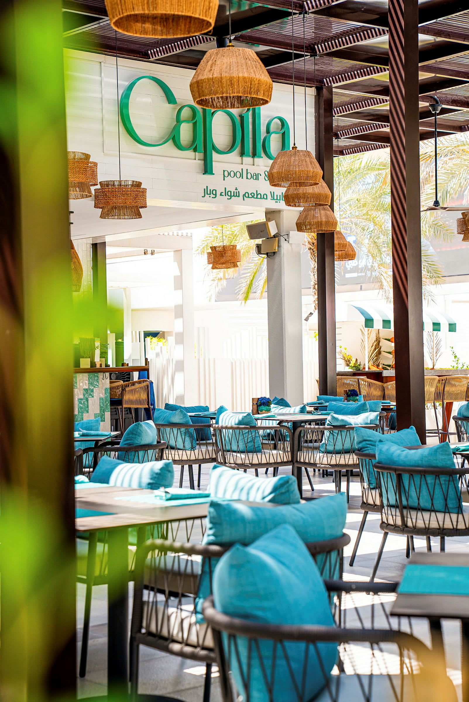 En utendørs bar og restaurant med turkise sittemøbler og dekorert med rottinglamper. Et skilt med teksten "Capila pool bar" er synlig i bakgrunnen. (AI-generert tekst)