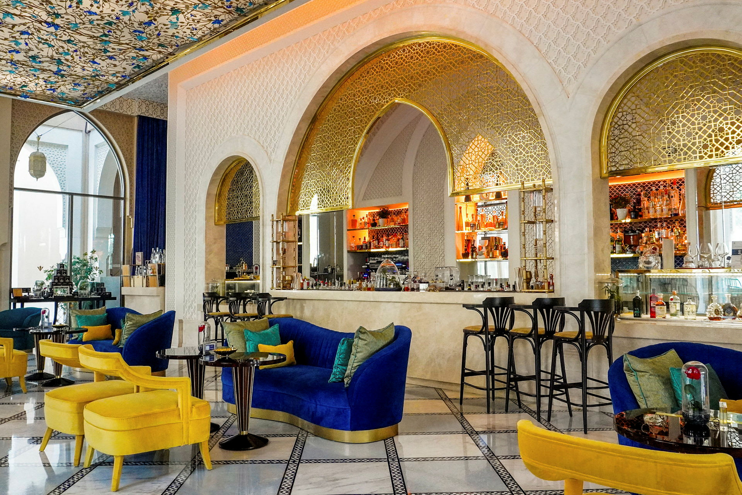 En elegant lounge med blå og gule møbler, et dekorativt tak og en bar med gullfargede detaljer. (AI-generert tekst)