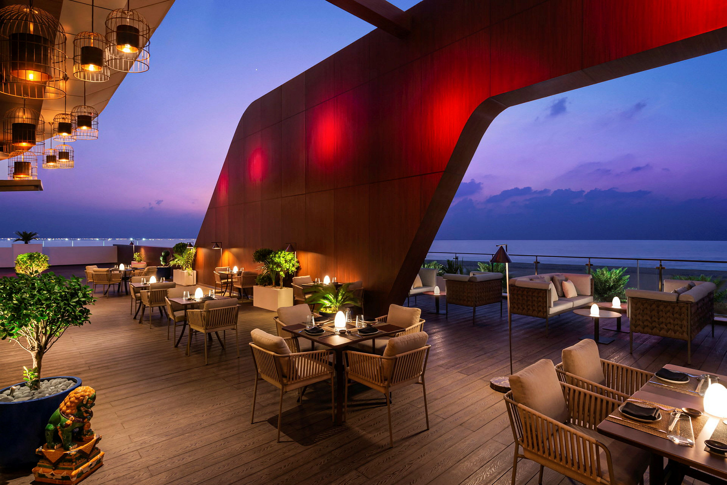 En moderne restaurantterrasse med bord og stoler, plassert ved havet på kveldstid med lamper og stilfull belysning. (AI-generert tekst)