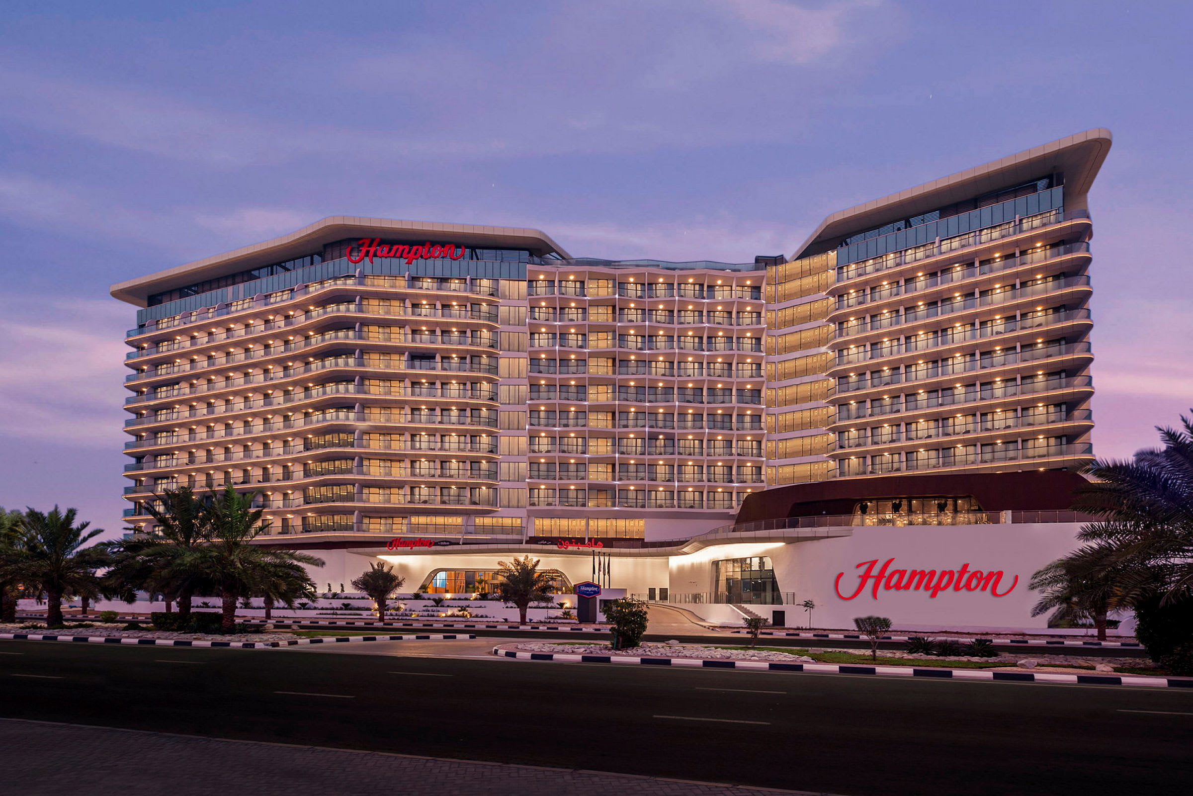 Et stort hotell med navnet "Hampton" vises i kveldslyset. Bygningen har flere etasjer med mange balkonger, og det er palmer foran. (AI-generert tekst)
