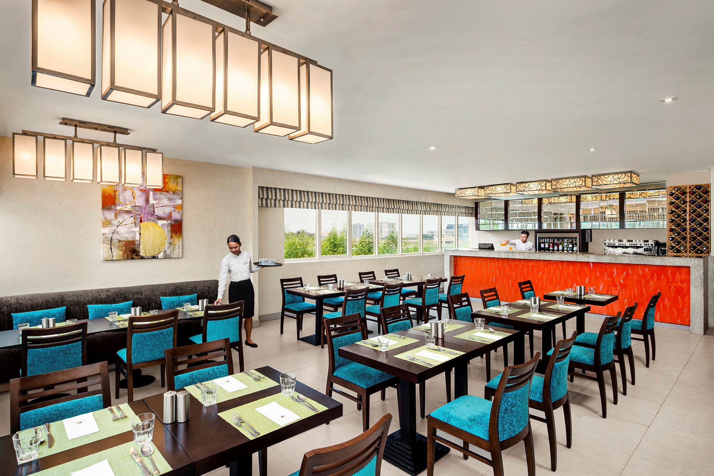 En elegant restaurant med bord og turkise stoler. Barområdet er innredet med en oransje panel, og store vinduer gir naturlig lys. (AI-generert tekst)