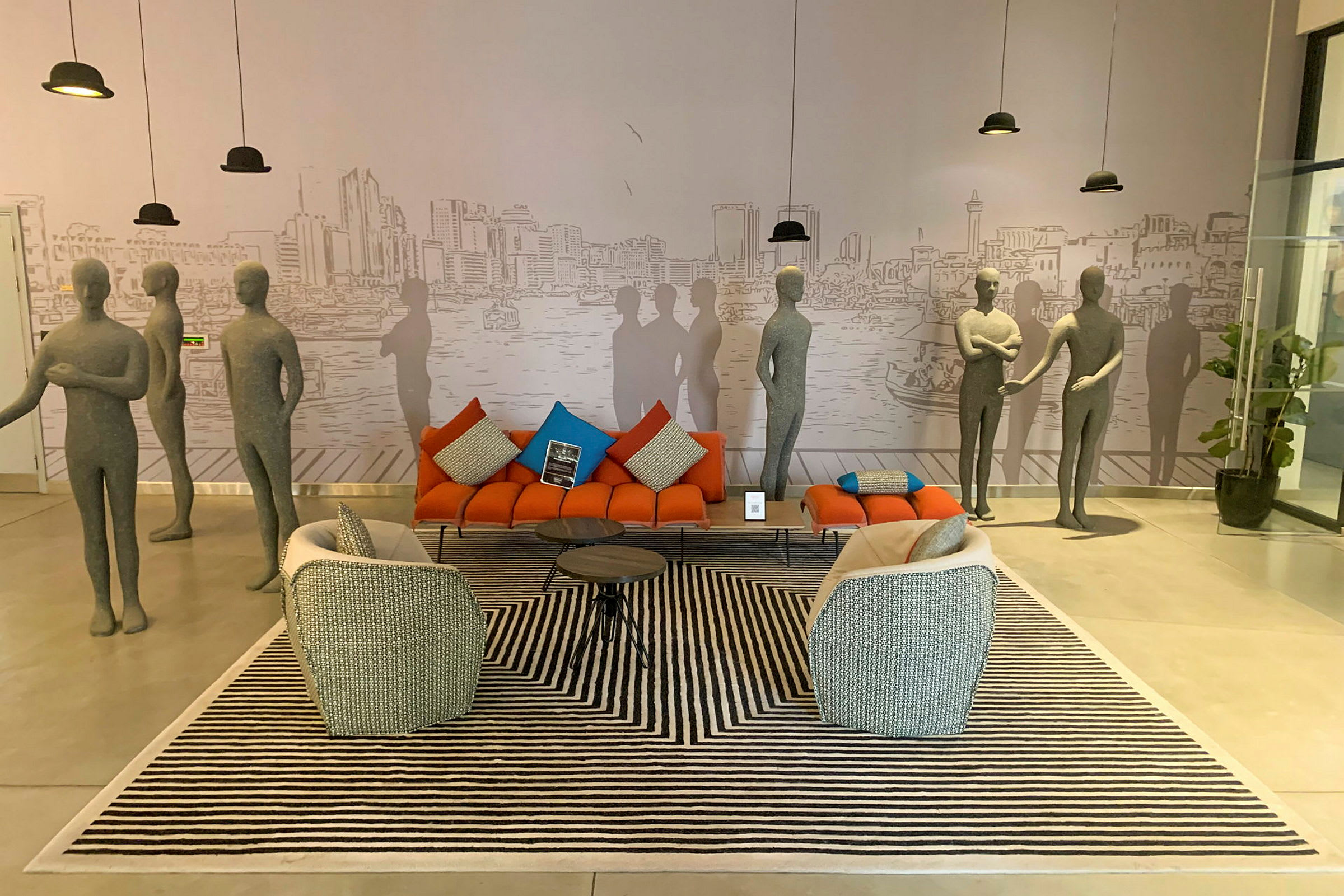 En moderne innredet stue med pent plasserte sofaer, lenestoler og et dekorativt bord på et stripete teppe. Det er flere grå mannequiner som står rundt i rommet, og et stort veggmaleri i bakgrunnen viser byens bygninger. (AI-generert tekst)