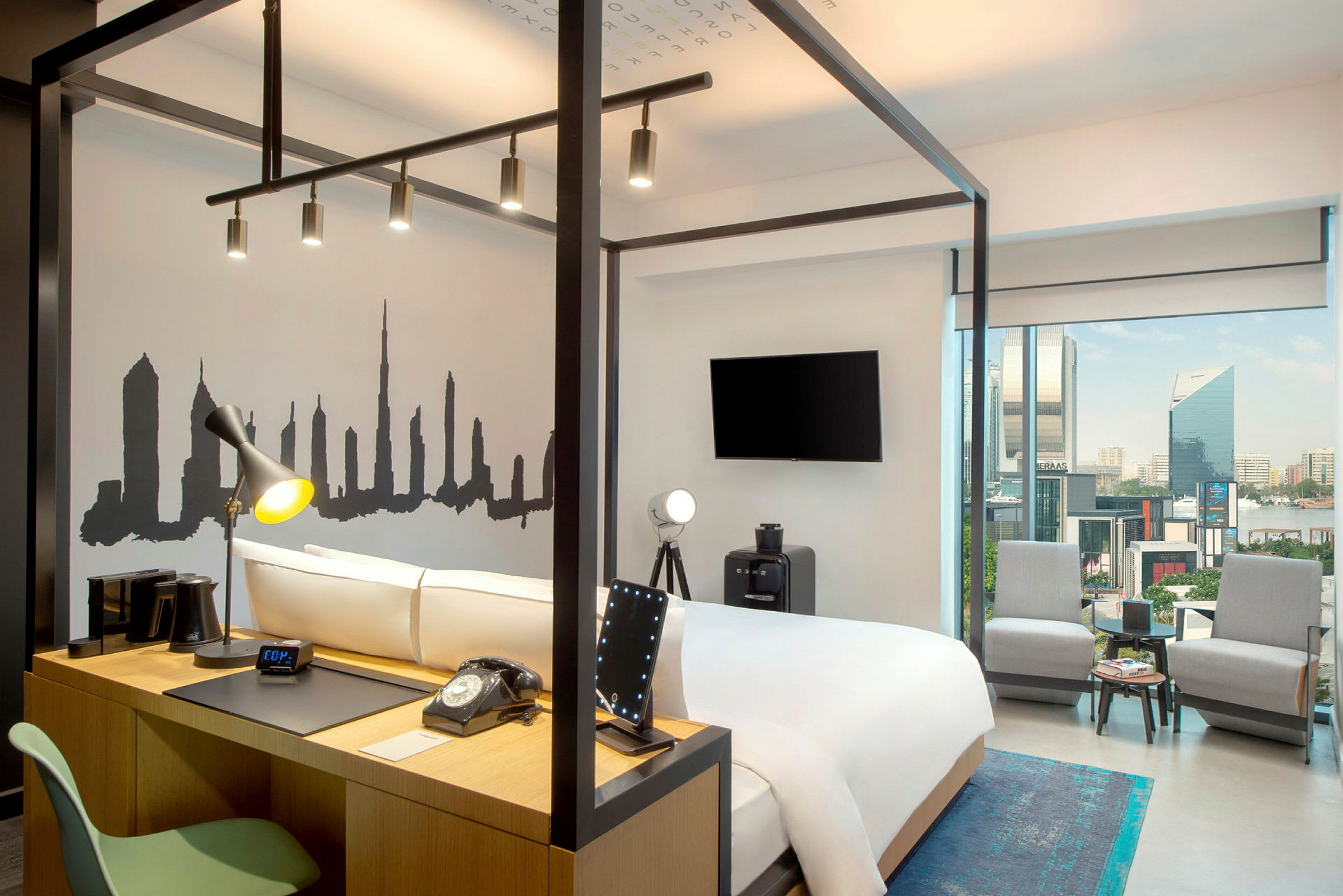Et moderne hotellrom med en seng omgitt av en stålkonstruksjon, en veggdekorasjon som viser en bysilhuett, og et skrivebord med en lampe og telefon. Store glassvinduer gir utsikt over byen. (AI-generert tekst)