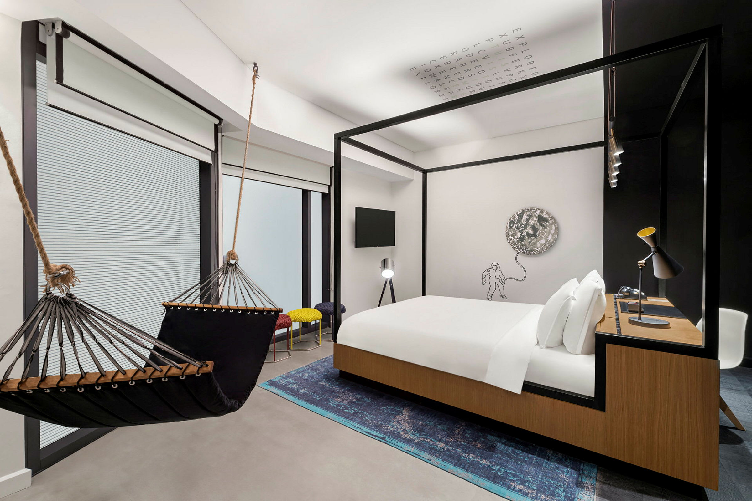 Et moderne hotellrom med en hengekøye, en dobbeltseng med tredetaljer, et skrivebord og en kunstnerisk veggdekorasjon. (AI-generert tekst)