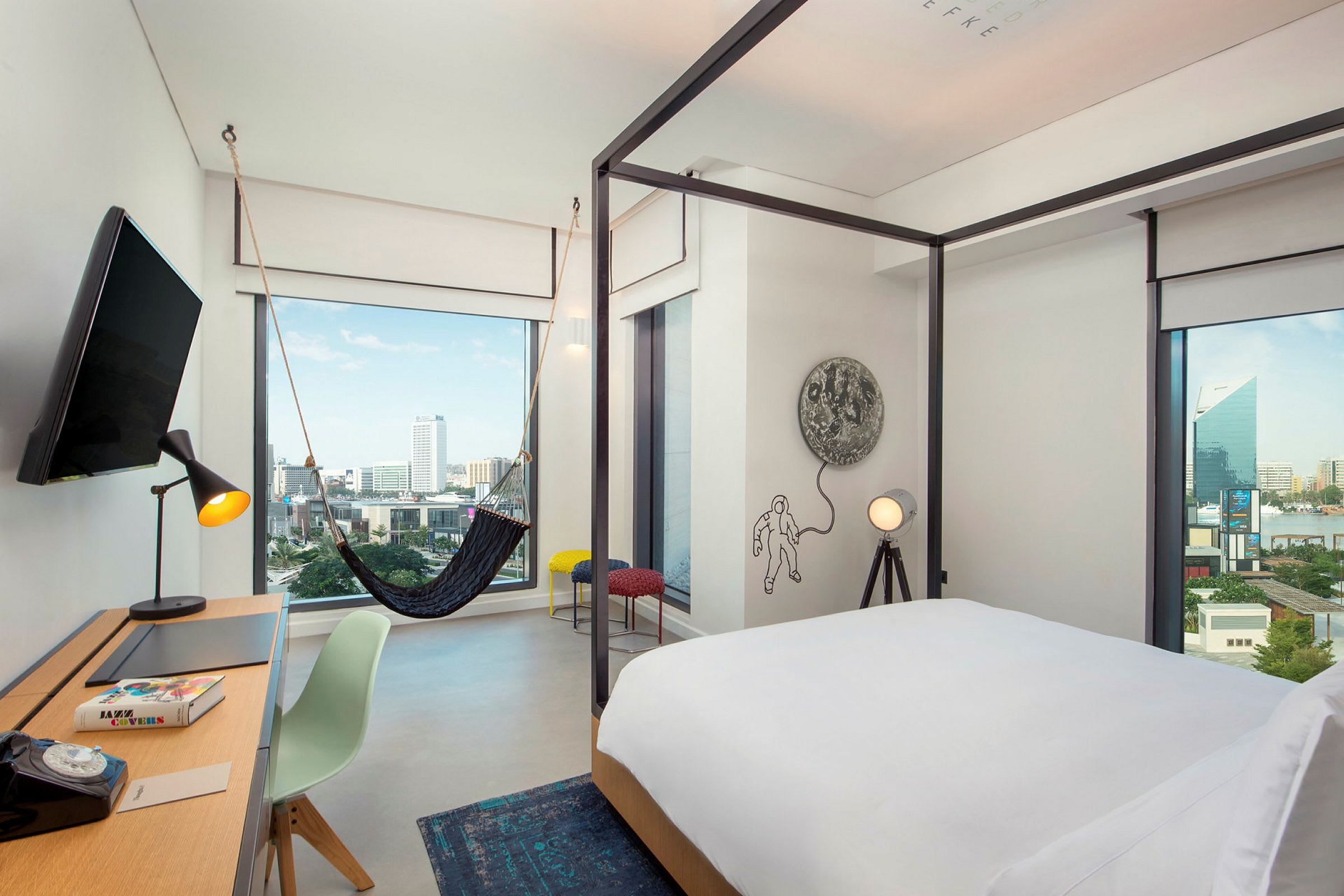 Et moderne hotellrom med en seng med hvitt sengetrekk, et skrivebord med en stol og en lampe, samt en hengekøye nær et stort vindu som viser utsikt over byen. (AI-generert tekst)