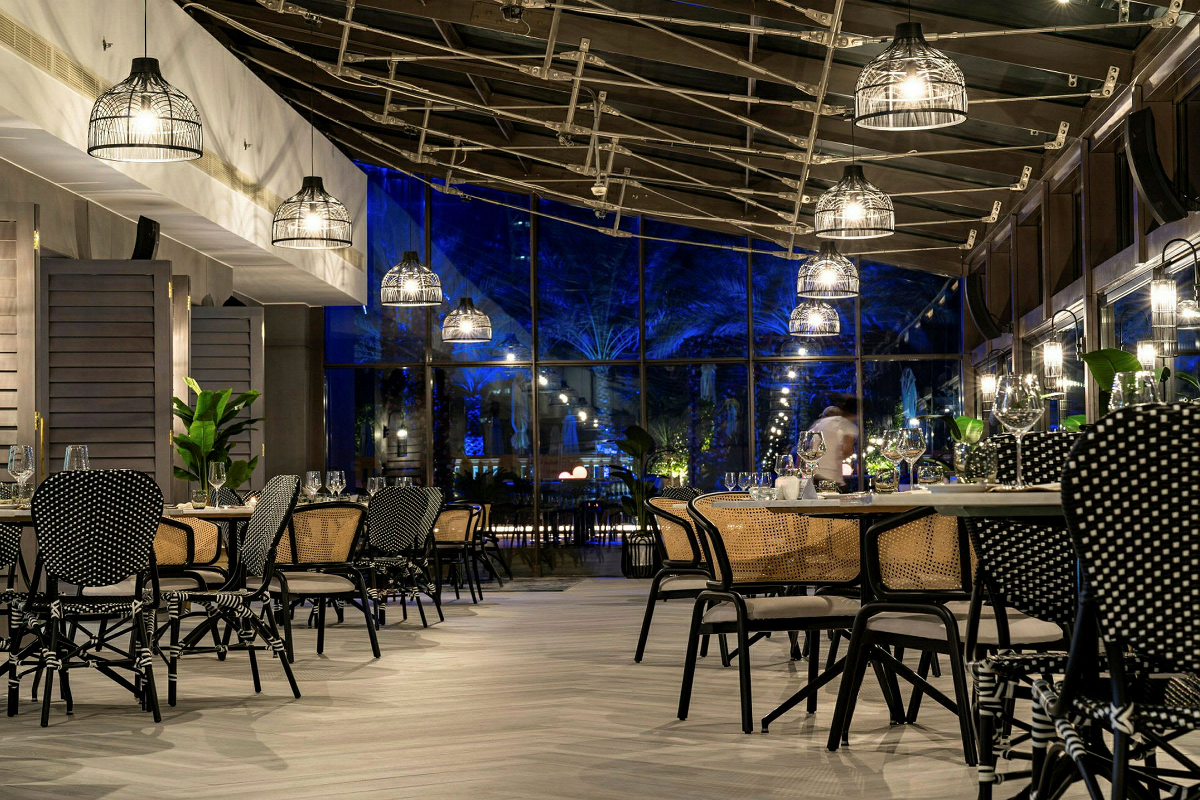 En restaurant med moderne møbler og taklamper, samt glassvegger som viser utendørs belysning om kvelden. (AI-generert tekst)