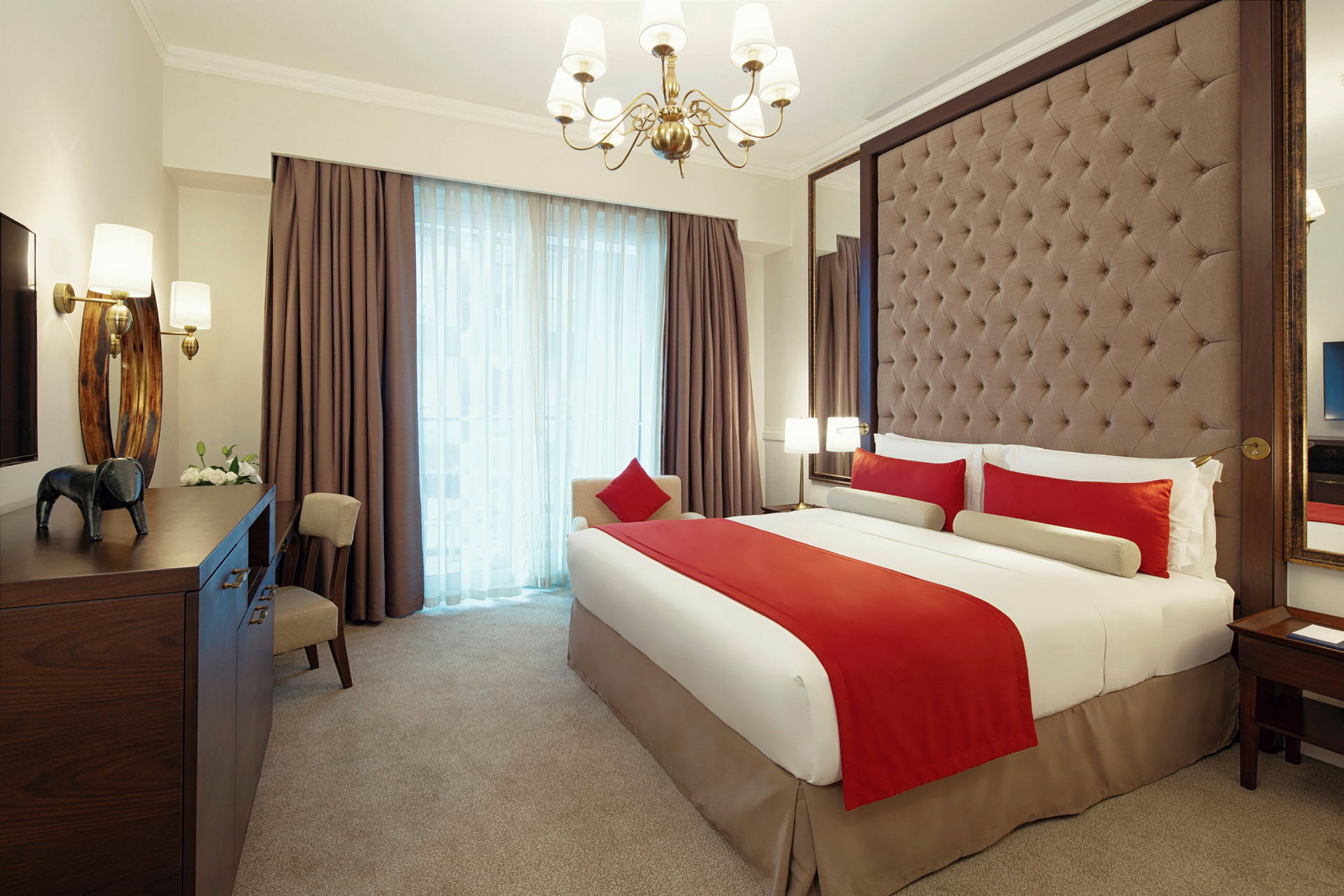 Et elegant innredet hotellrom med en stor seng kledd i hvitt og rødt sengetøy. Rommet har en polstret sengegavl, en lysekrone og store vinduer med gardiner. (AI-generert tekst)