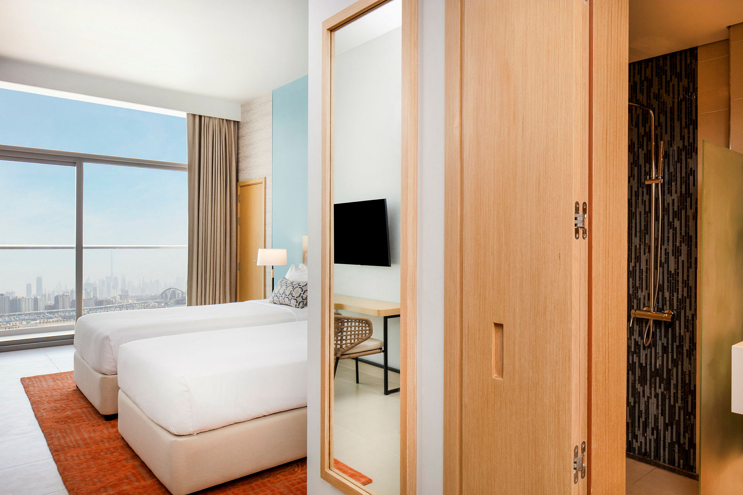 Et moderne hotellrom med to enkeltsenger som står nær et stort vindu med utsikt over en bysilhuett. Et bad syns delvis til høyre. (AI-generert tekst)