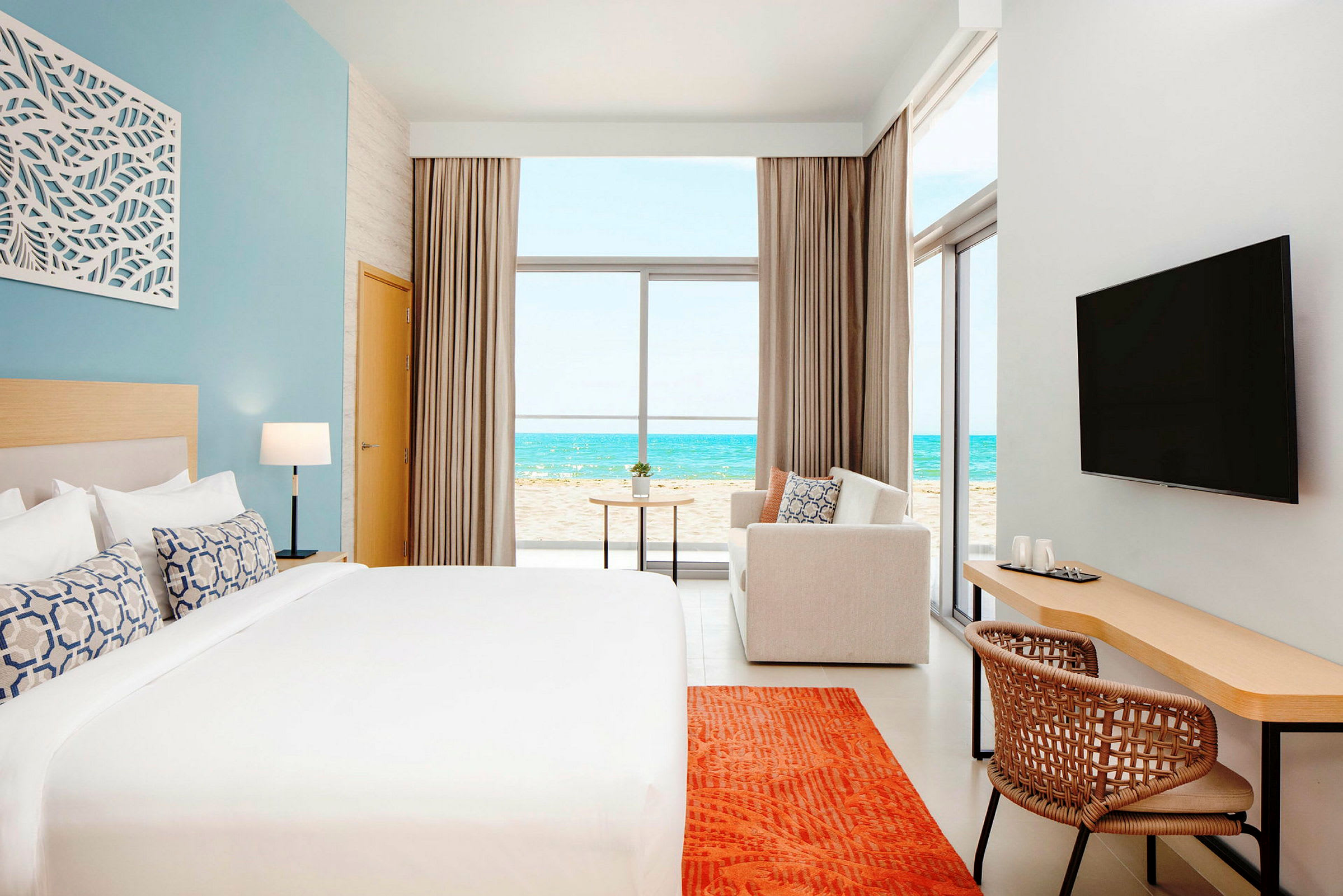 Et moderne hotellrom med en stor seng, en sofa, skrivebord og en TV. Utsikten fra rommet viser en strand og havet. (AI-generert tekst)