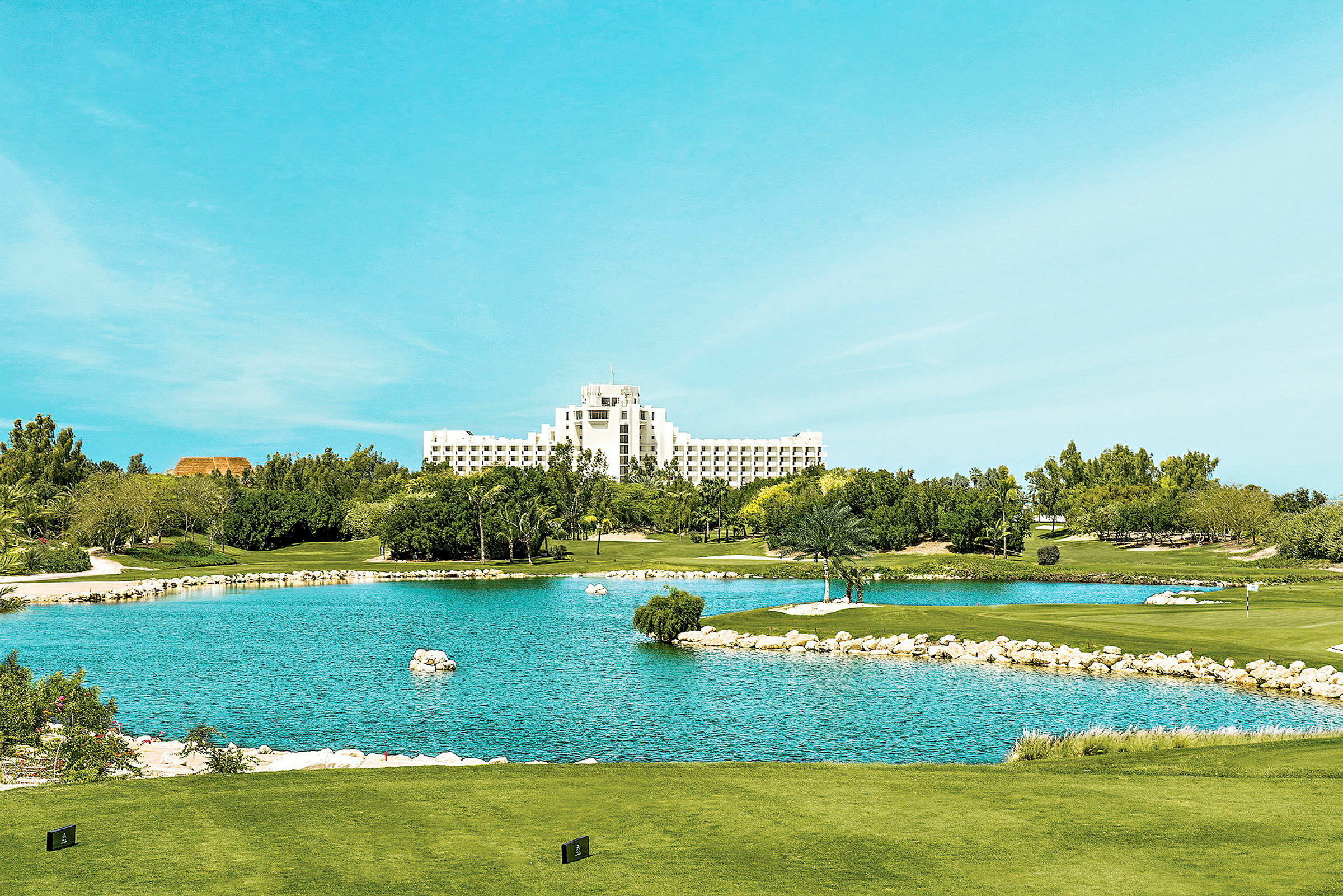 Utsikt over en golfbane med en dam i forgrunnen og et stort hvitt hotell i bakgrunnen. (AI-generert tekst)
