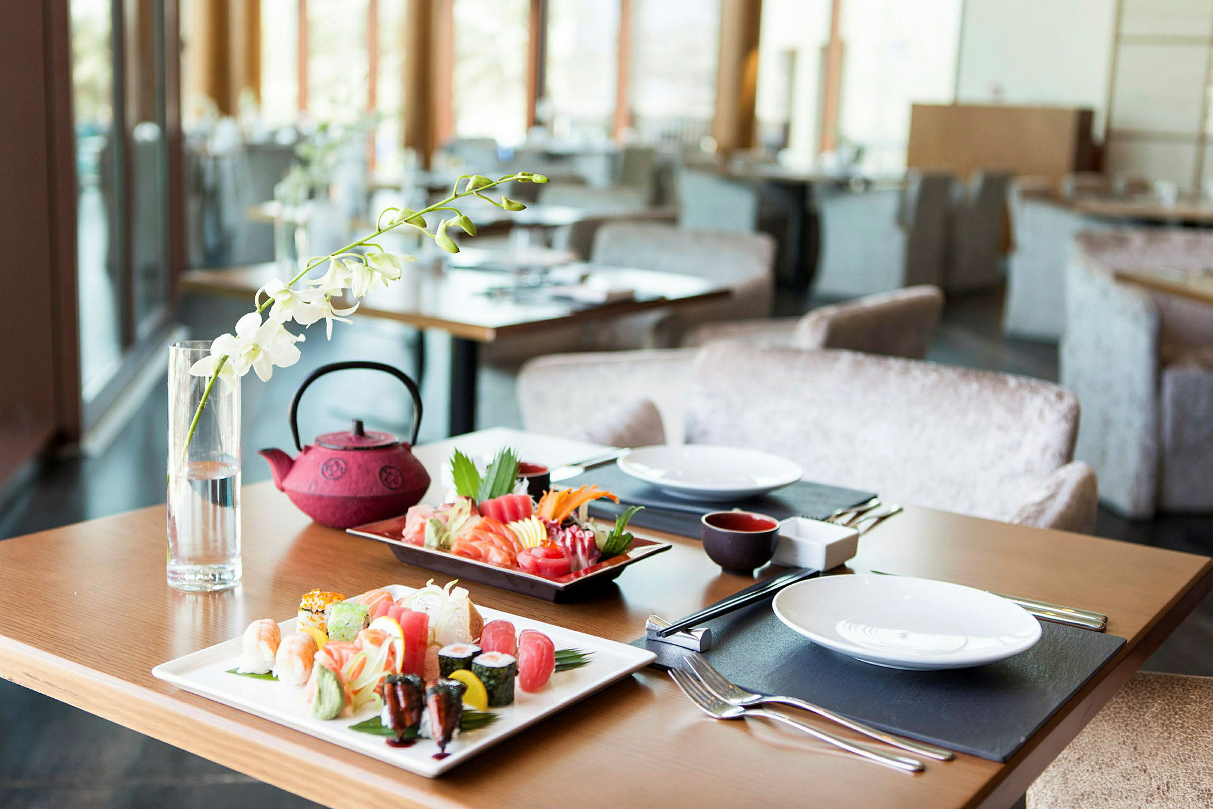 Et bord på en restaurant er dekket med sushi på fat, en rød tekanne og et glass med en hvit blomst som dekorasjon. (AI-generert tekst)