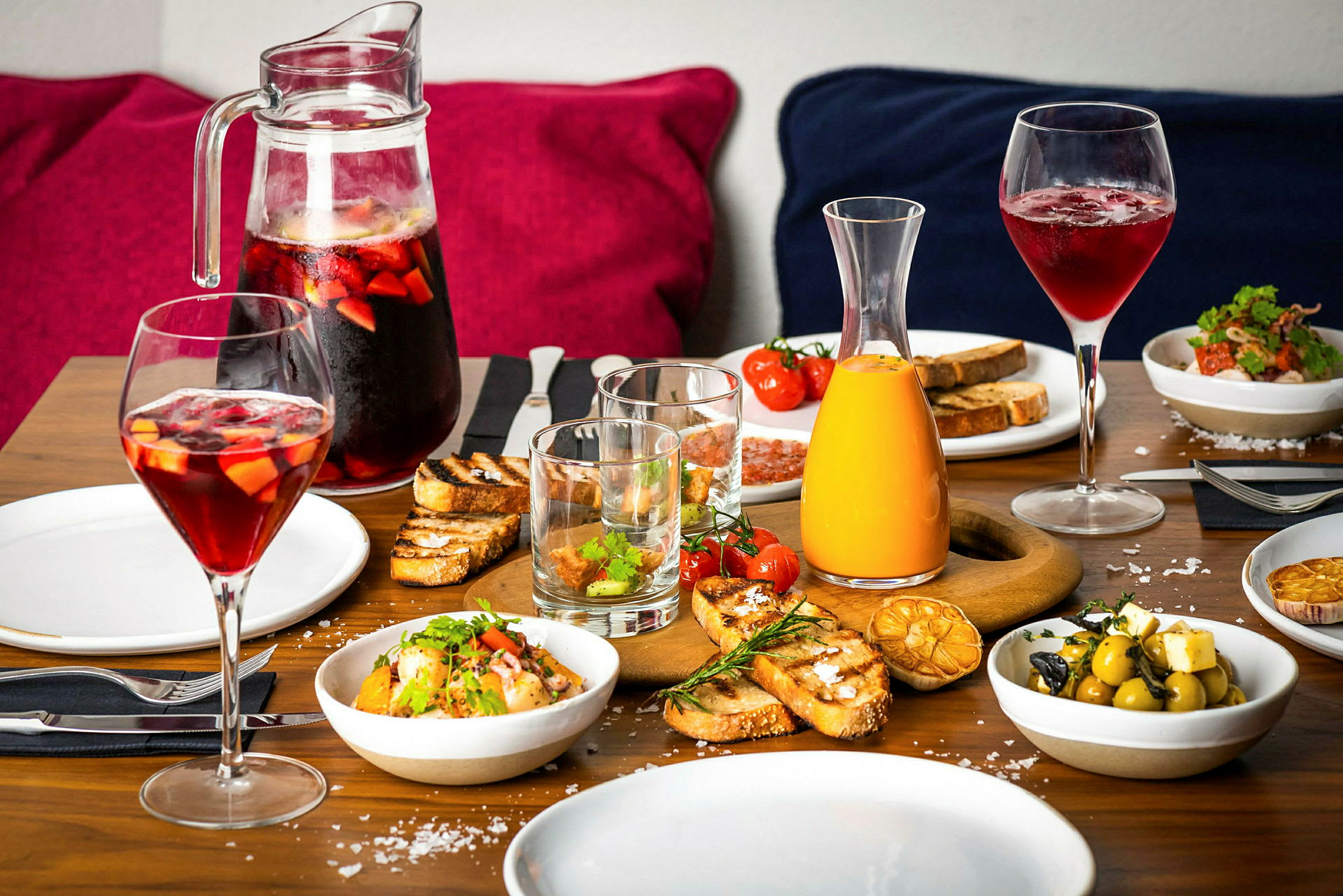 Et bord dekket med mat og drikke, inkludert salat, brød, oliven og karafler med juice og sangria. (AI-generert tekst)