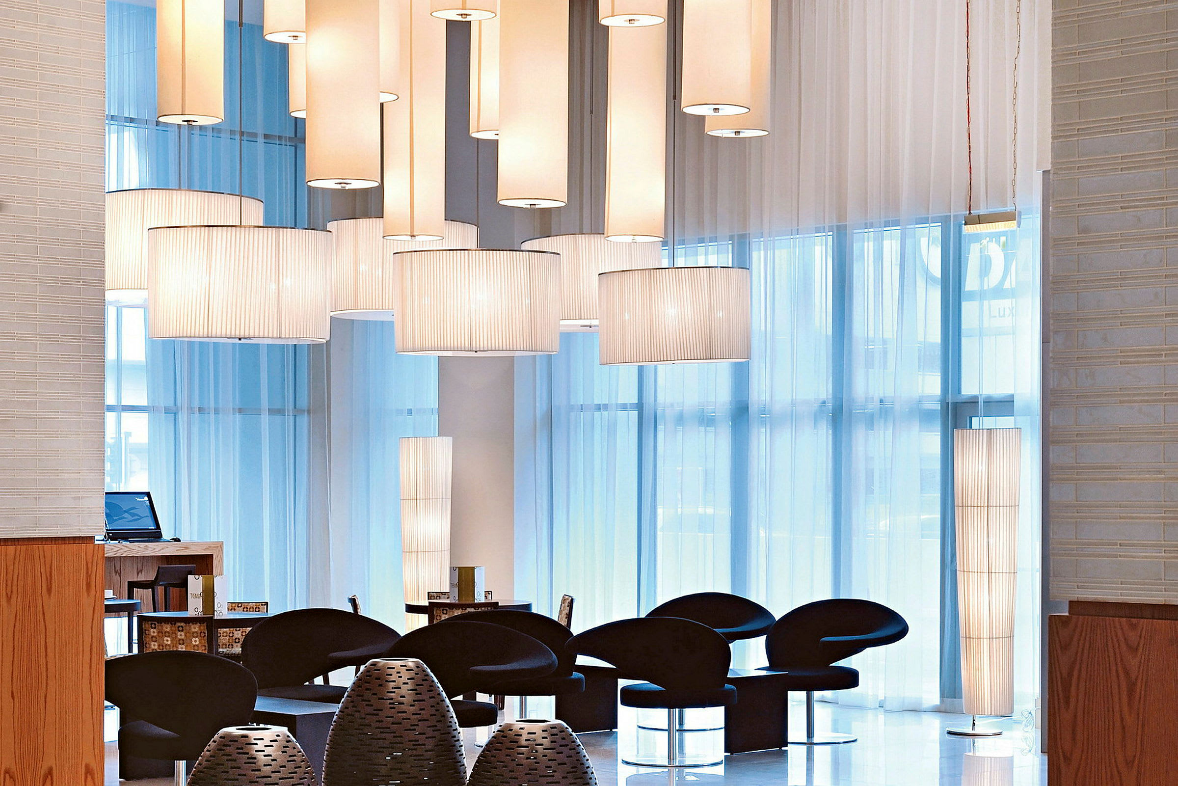Interiørutsikt med en moderne lounge som inneholder svarte stoler, hengende lamper og store vinduer som slipper inn dagslys. (AI-generert tekst)