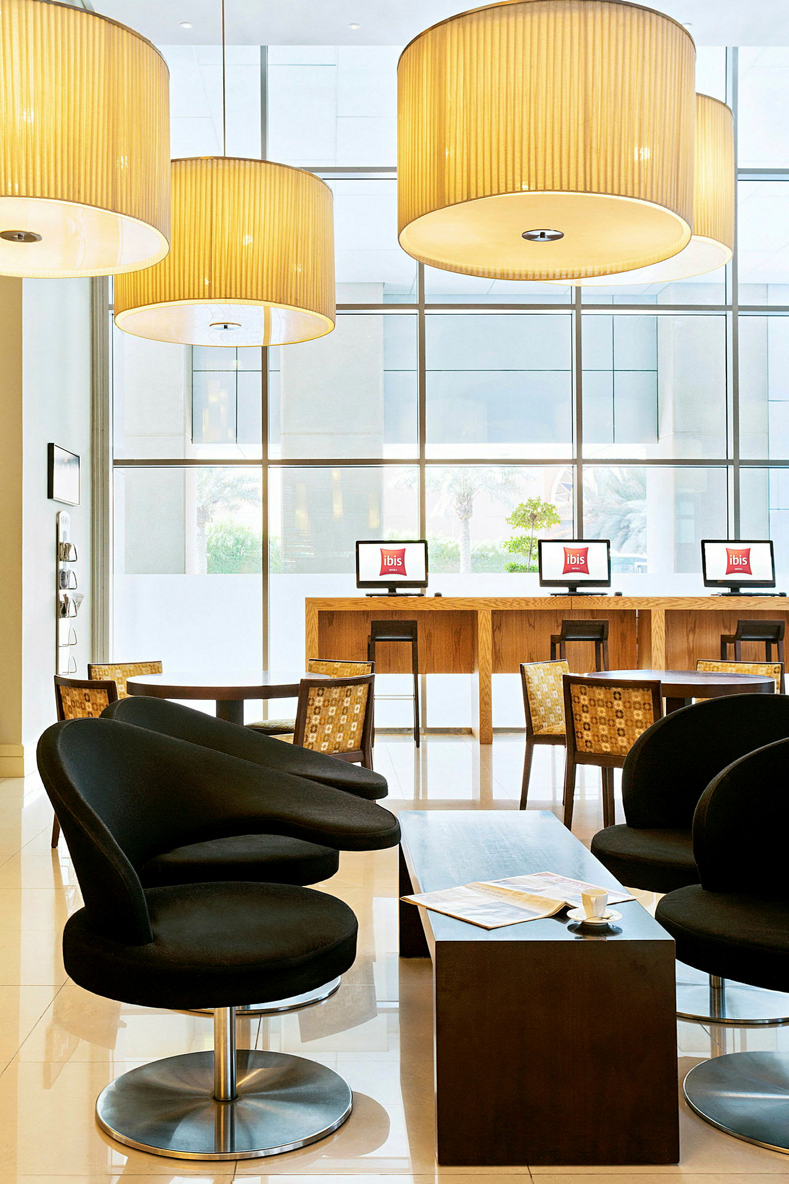 Et hotellobbyområde med svarte lenestoler, et lavt bord og datamaskiner på en bardisklignende benk foran store vinduer. (AI-generert tekst)